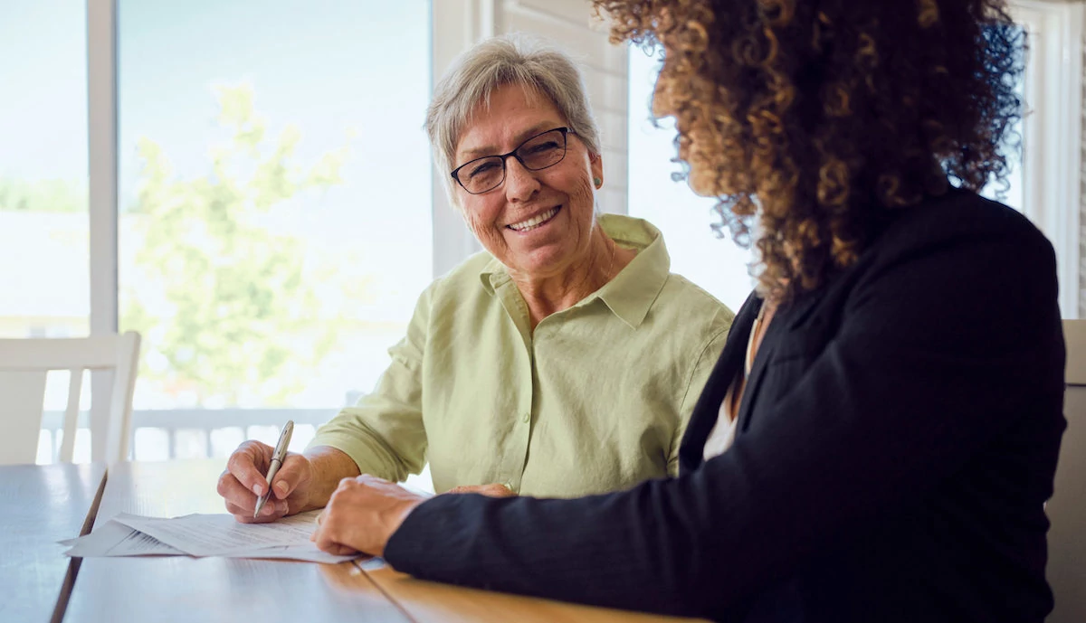 Two women seated at a table with papers between them. One woman holds a pen to the paper while smiling at the camera.