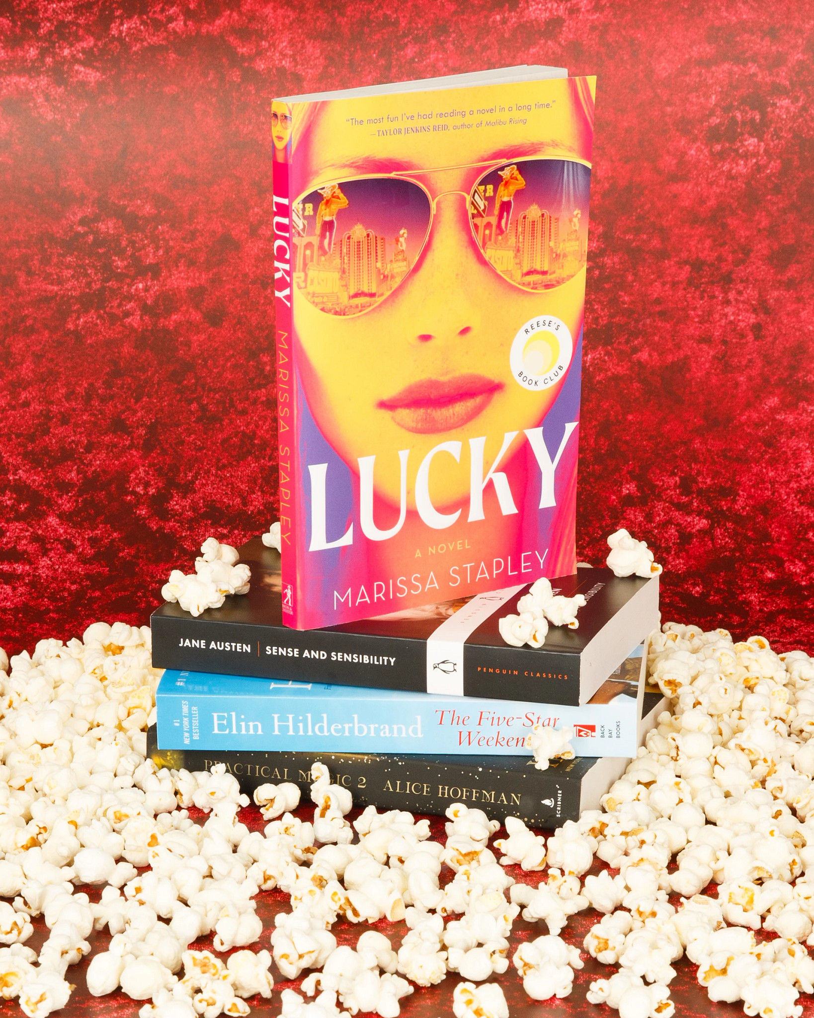 A brightly colored book titled “Lucky” by Marissa Stapley is displayed on top of a stack of novels surrounded by popcorn against a red textured background