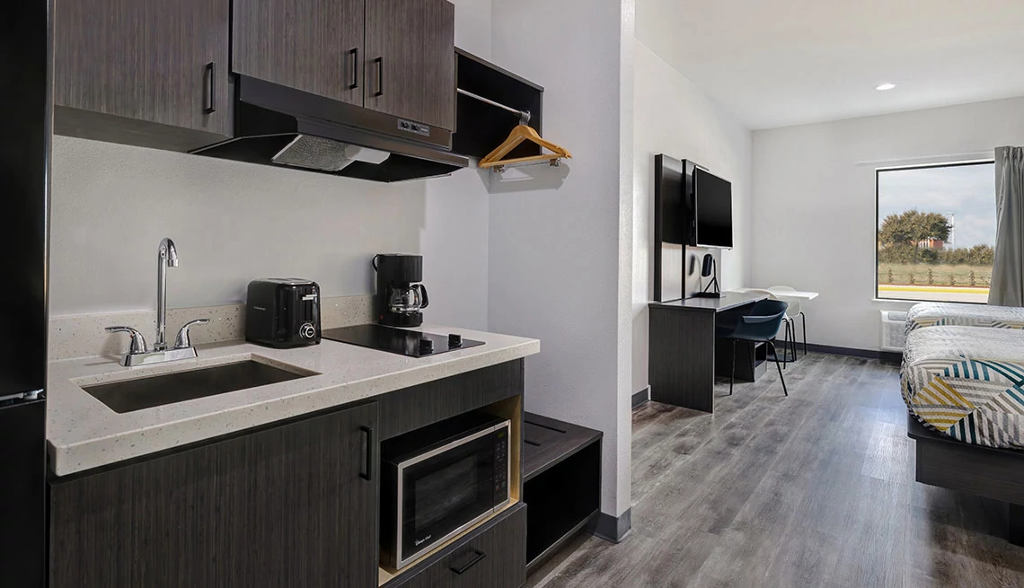 Studio 6 hotel room with gray wood floors large window with open curtain, end of bed showing, desk, flat screen tv mounted to the wall, small kitchenette with sink, small stove top, microwave, coffee maker, toaster, cabinets overhead, small closet space with wood hangers showing