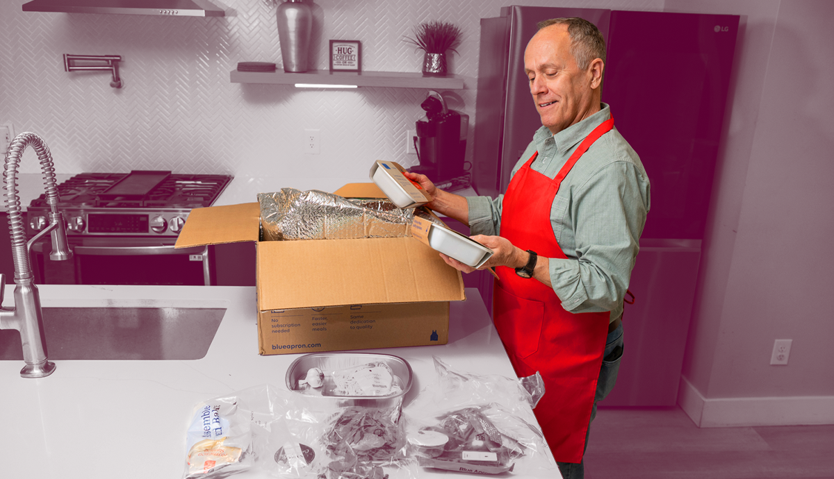 A person holding a Blue Apron recipe card with the meal kit box and ingredients spread out on the counter.