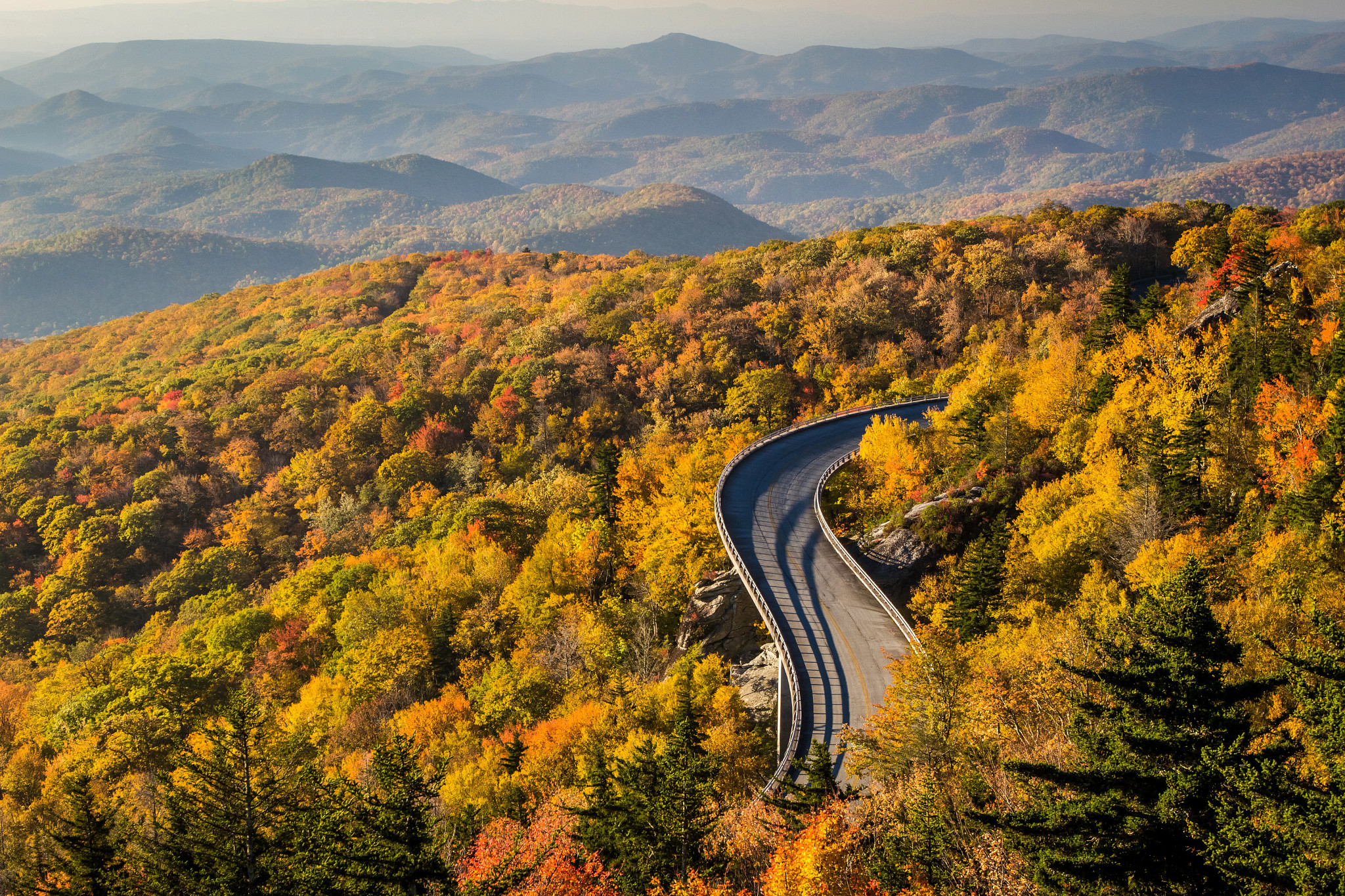 Panoramic mountain landscape with a winding road surrounded by colorful autumn trees in shades of gold, orange, and green