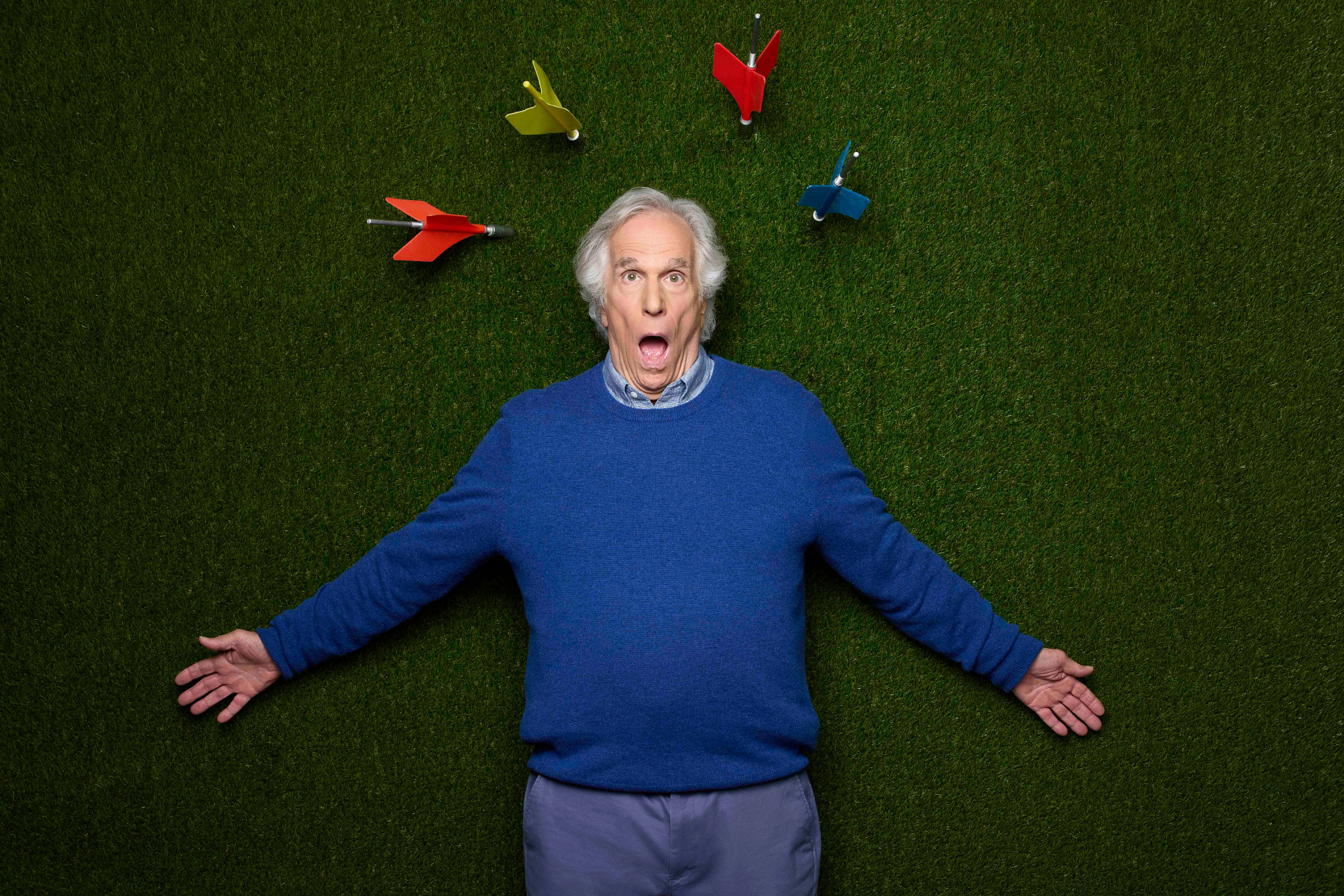 Henry Winkler making a comical surprised face while lying on an astroturf background with his head surrounded by lawn darts