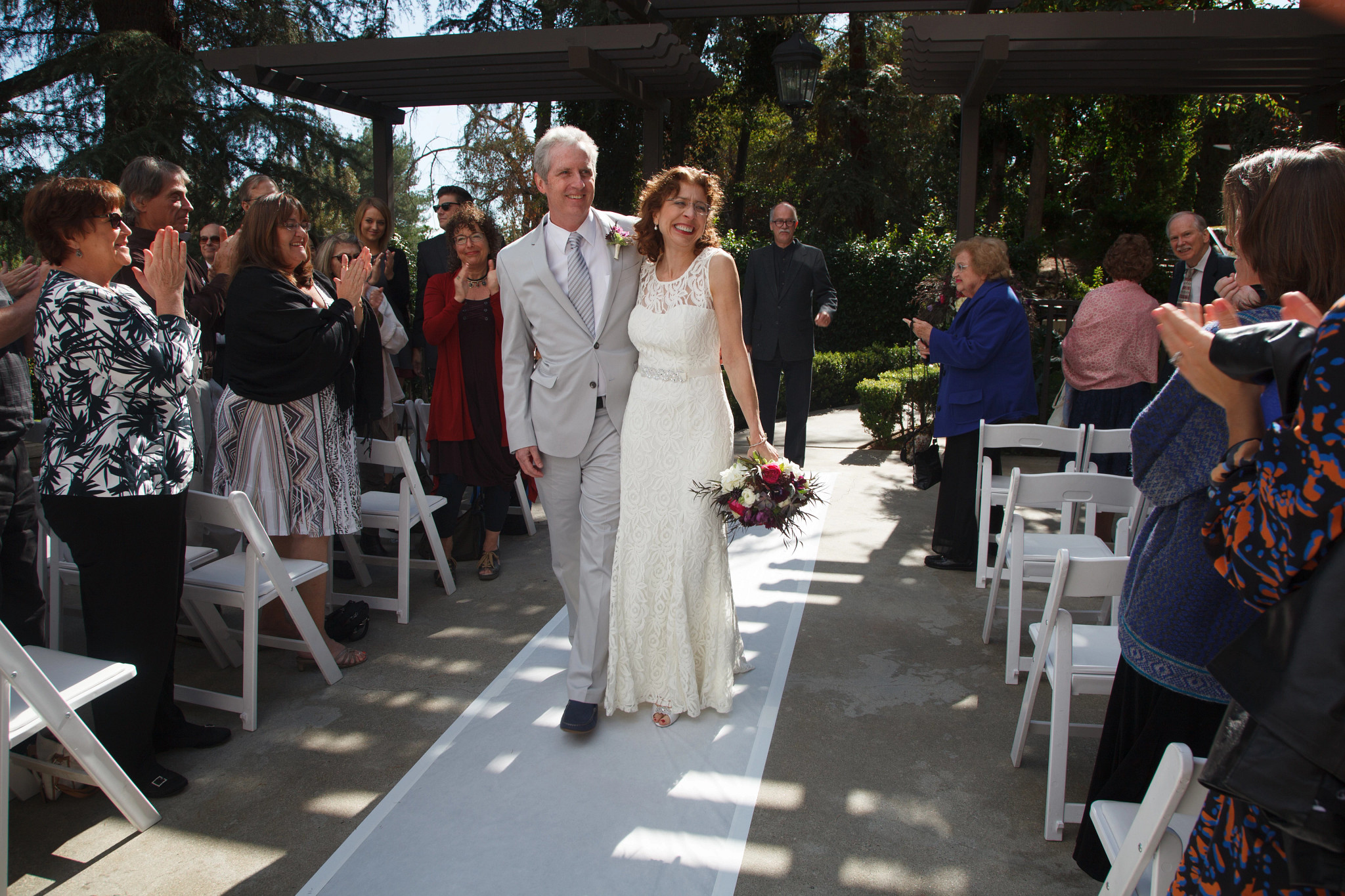 Laura Sturza and her husband Tom walking down an outdoor wedding aisle surrounded by applauding guests standing near white chairs