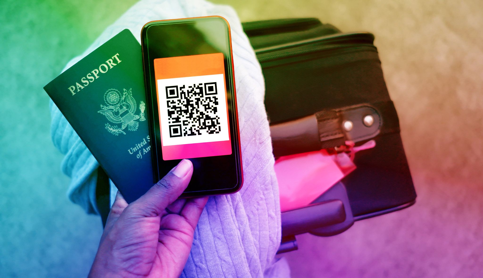 Una mano sostiene un pasaporte estadounidense y un celular cuya pantalla muestra un código qr. Atrás se ve un sweater doblado arriba de una maleta negra con una etiqueta roja.