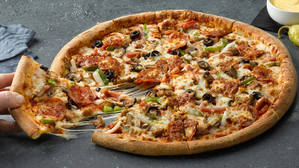 large supreme pizza laying on a slate background with a male hand taking a slice of the pizza, garlic sauce, and pepperoncini in the background