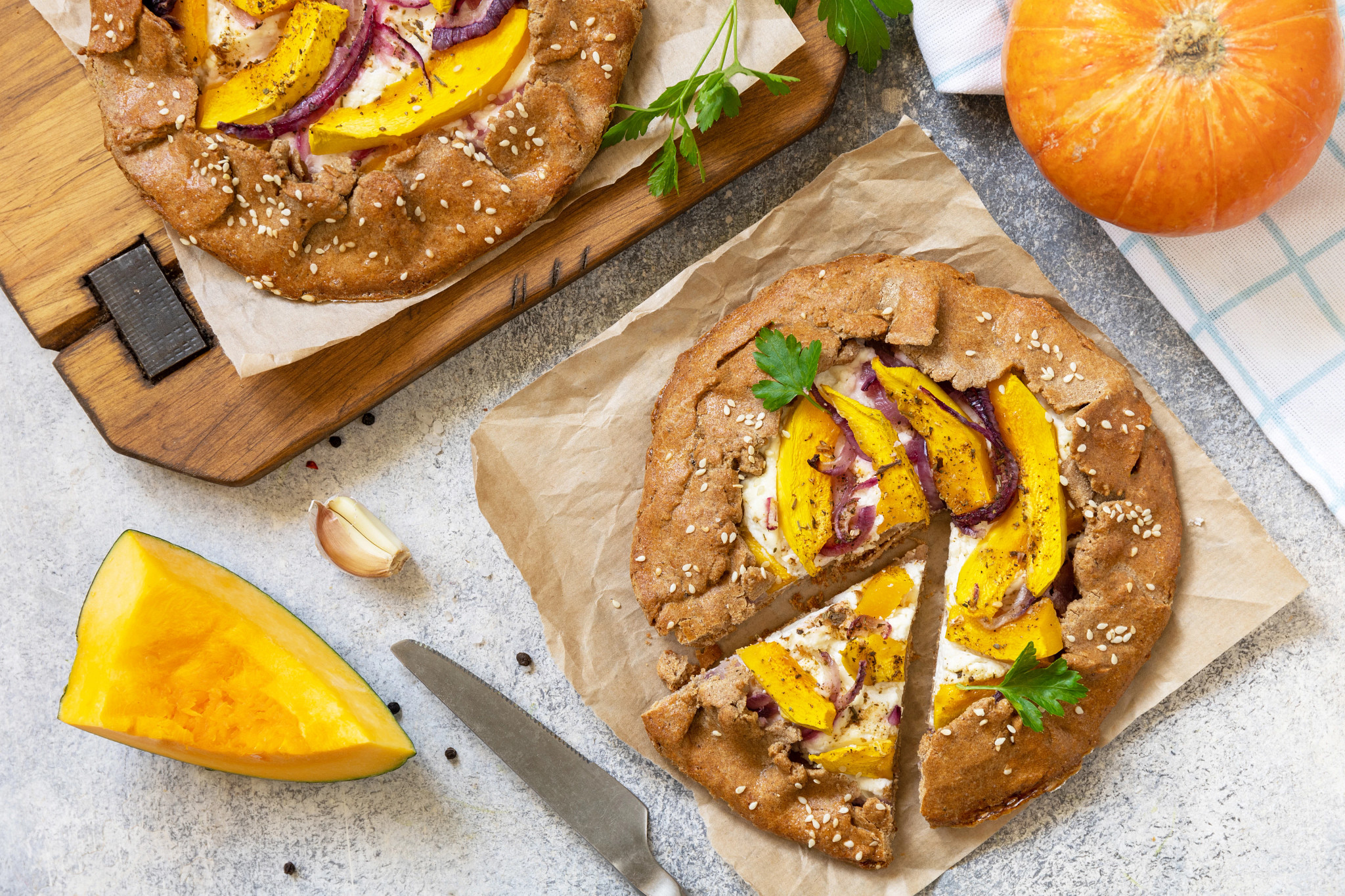 Overhead view of a rustic savory pumpkin galette made with pumpkin slices, ricotta cheese, red onions, and sesame seeds on a parchment-lined baking sheet. A sliced wedge reveals the filling, while fresh pumpkin and garlic rest nearby