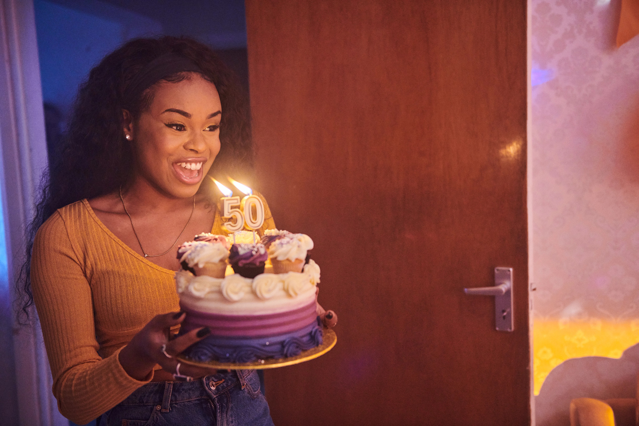 A smiling woman holding a birthday cake decorated with cupcakes and lit candles shaped like the number 50.