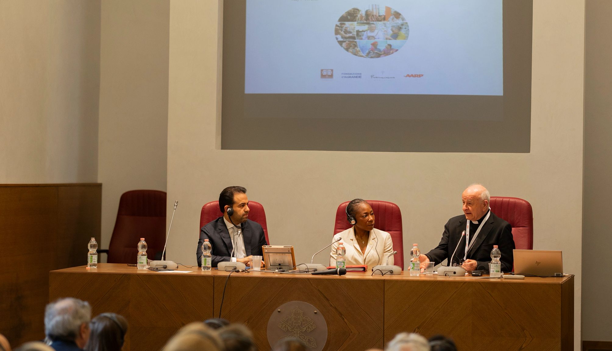 AARP and the Vatican Open Historic Summit three people sit at a table, facing a crowd of conference attendees