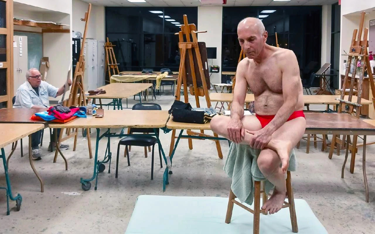 a photo shows Harold Kahn, wearing a speedo, atop a stool posing for a life drawing class at his Florida retirement community’s arts club