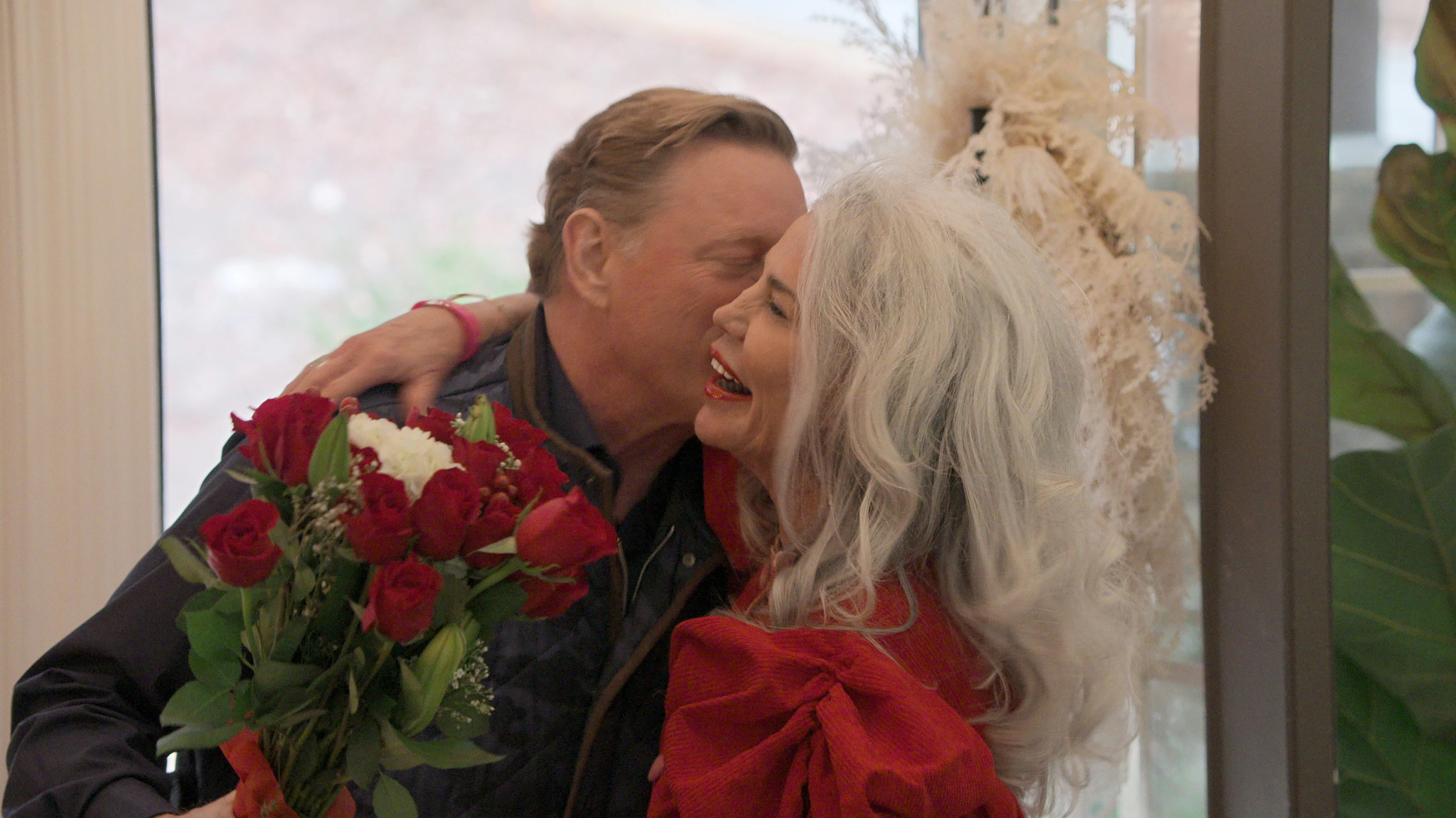 a man, holding a bouquet of flowers, hugs a woman and kisses her on the cheek