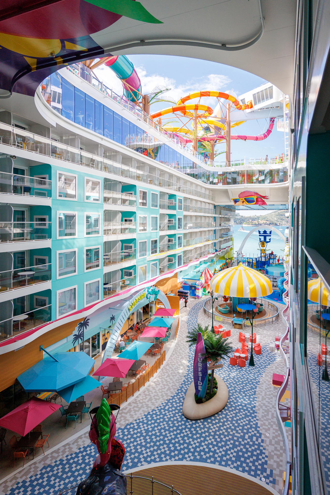Interior view of a modern cruise ship's Surfside neighborhood featuring colorful waterslides overhead, turquoise balcony staterooms, outdoor seating, umbrellas, and family-friendly recreation spaces
