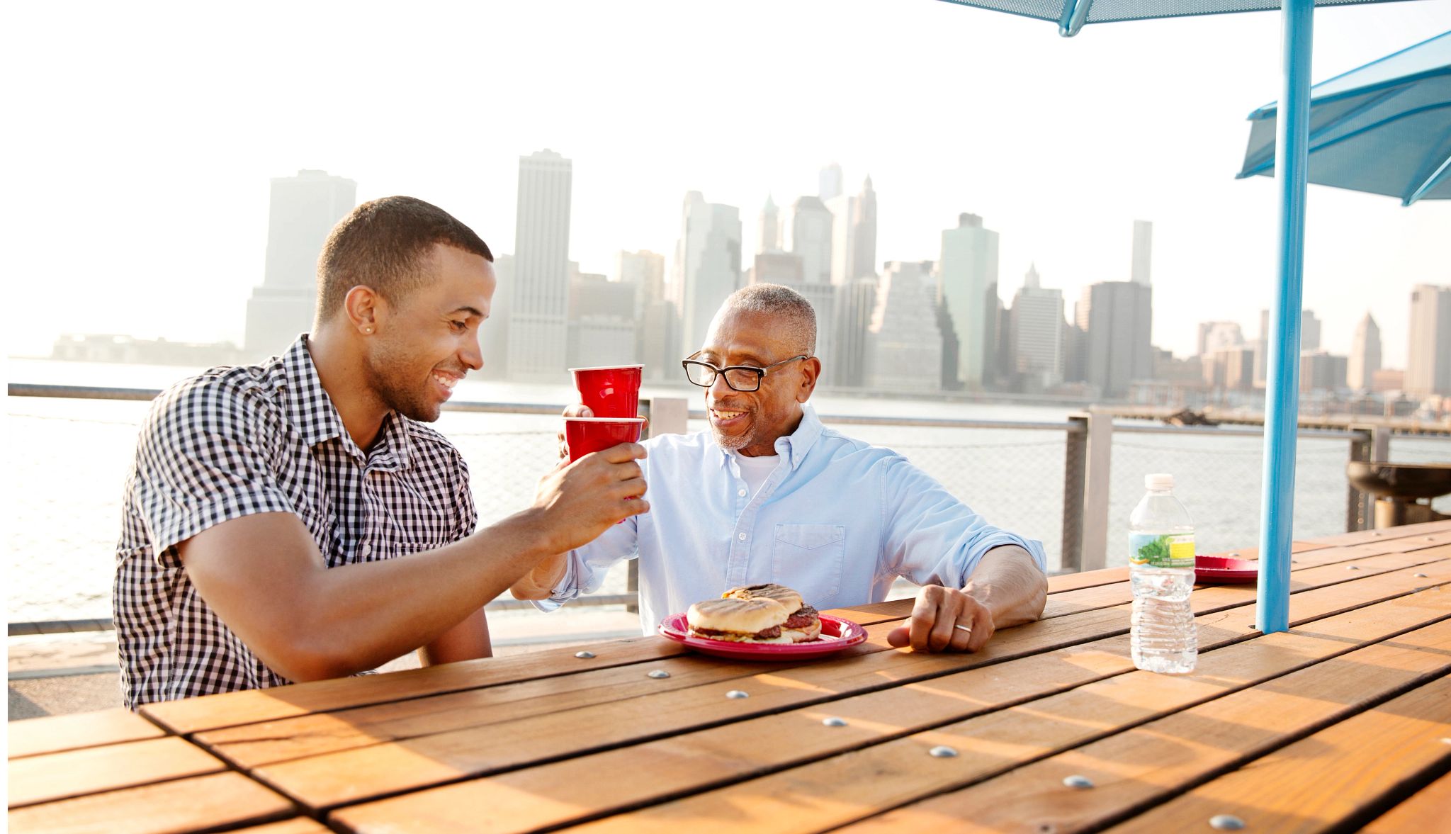 Father's Day Deals A photo shows a father and son celebrating at a picnic table with food and drink. A city skyline is behind them.