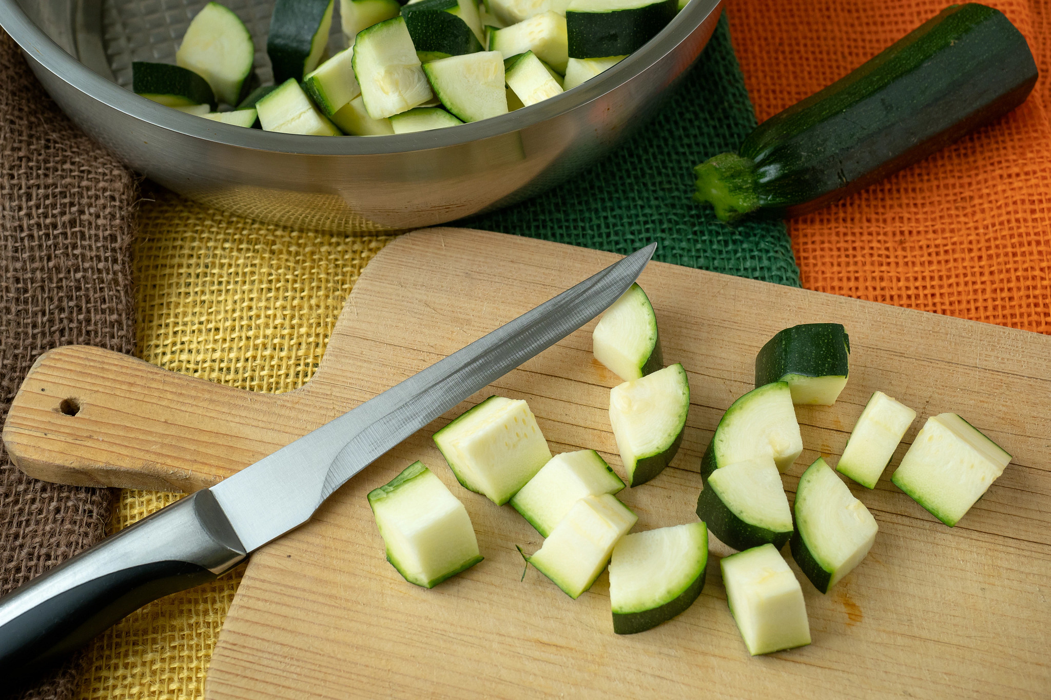 Fresh zucchini pieces chopped on a wooden cutting board with a kitchen knife and bowl of vegetables nearby