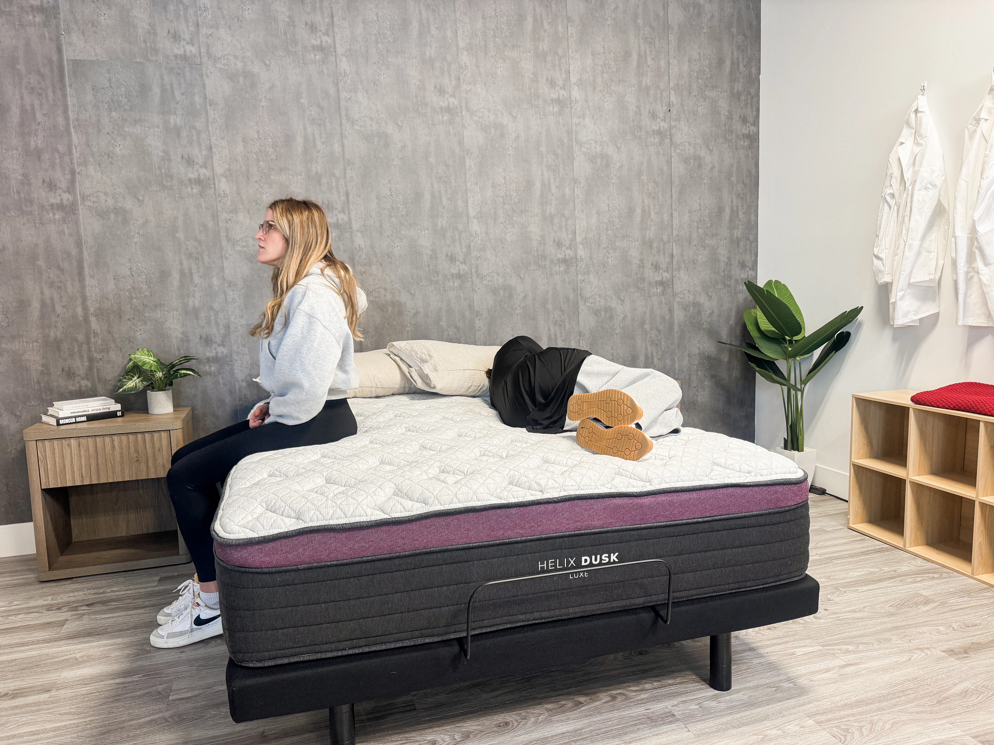 One person sits on the edge of the Helix Dusk Luxe mattress as another lies on the other side