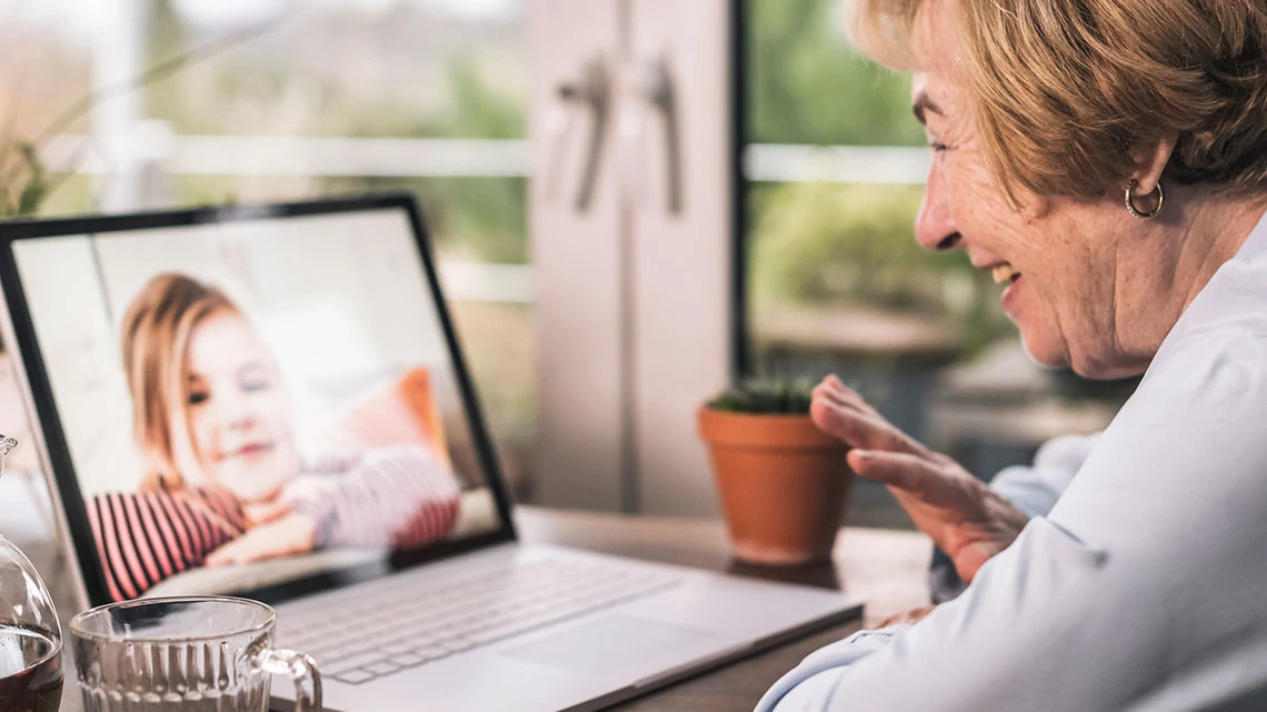 grandmother waving and smiling at laptop computer on kitchen table with small granddaughter on screen