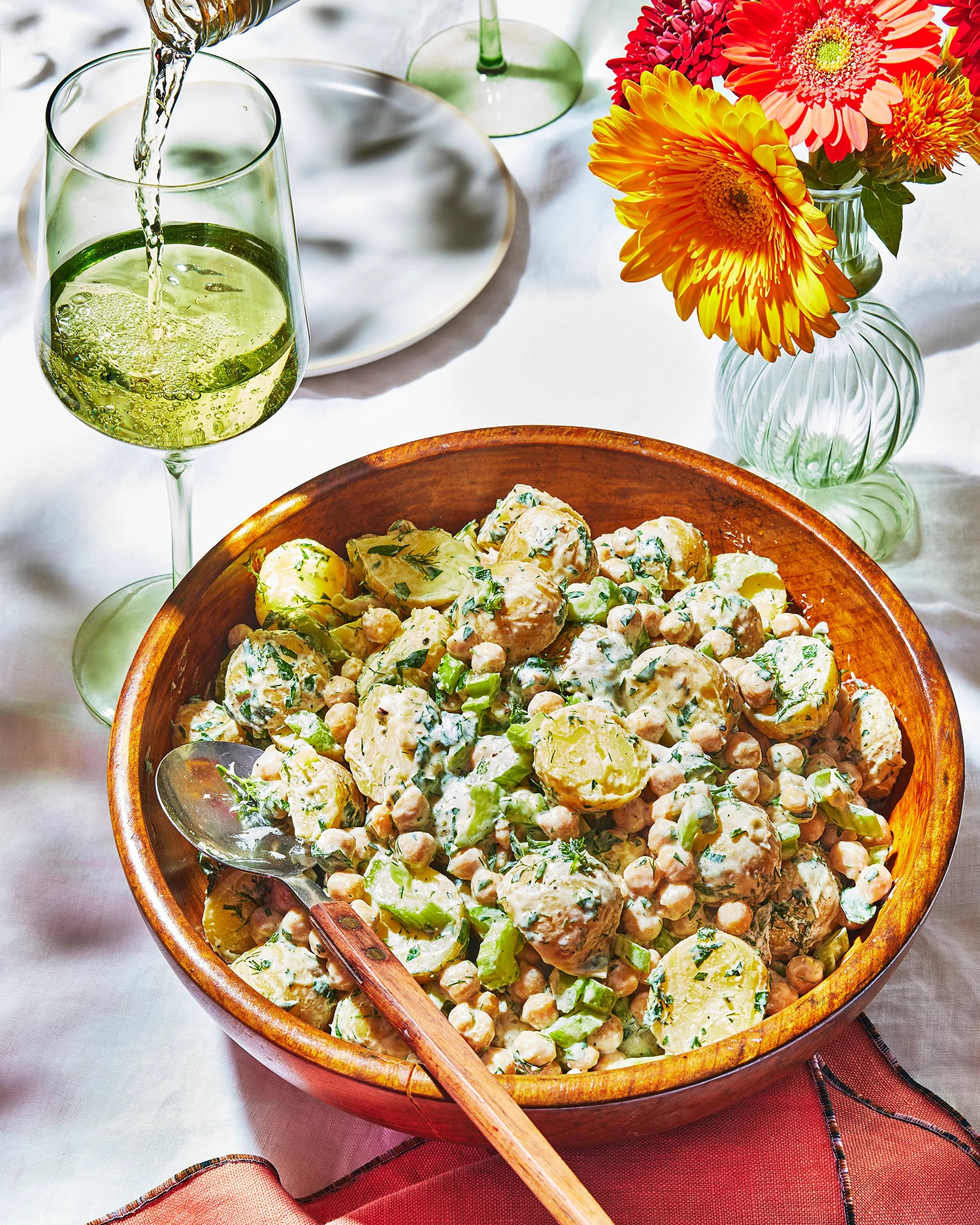 A vibrant bowl of potato chickpea salad with lemon yogurt dressing served on a bright table, alongside a glass of white wine and colorful flowers