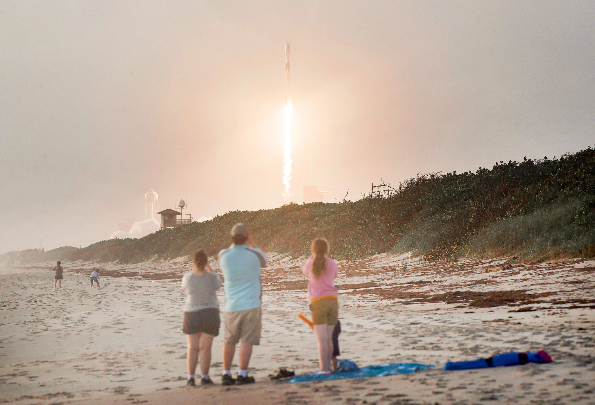 a rocket launches in the distance as spectators watch in the foreground on a beach