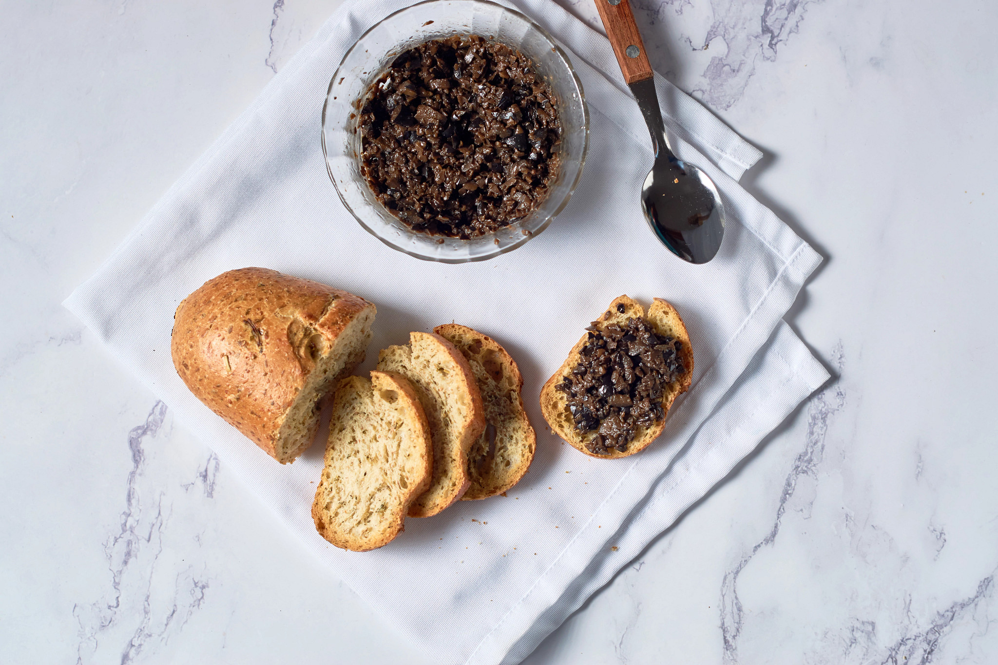 Bowl of eggplant and olive tapenade spread served alongside sliced artisan bread on a white cloth