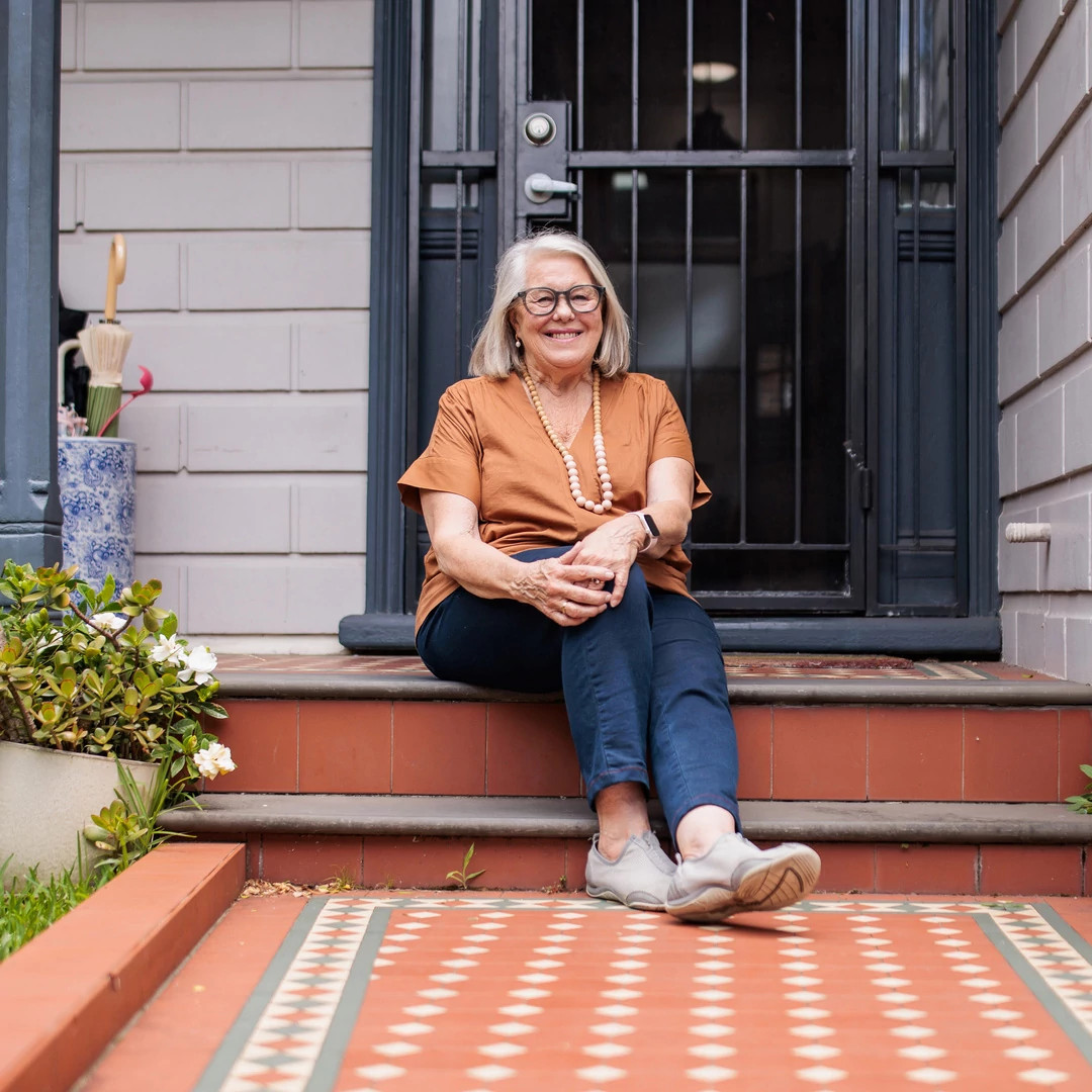 A smiling older woman in an orange top is sitting contentedly on her home’s front steps.