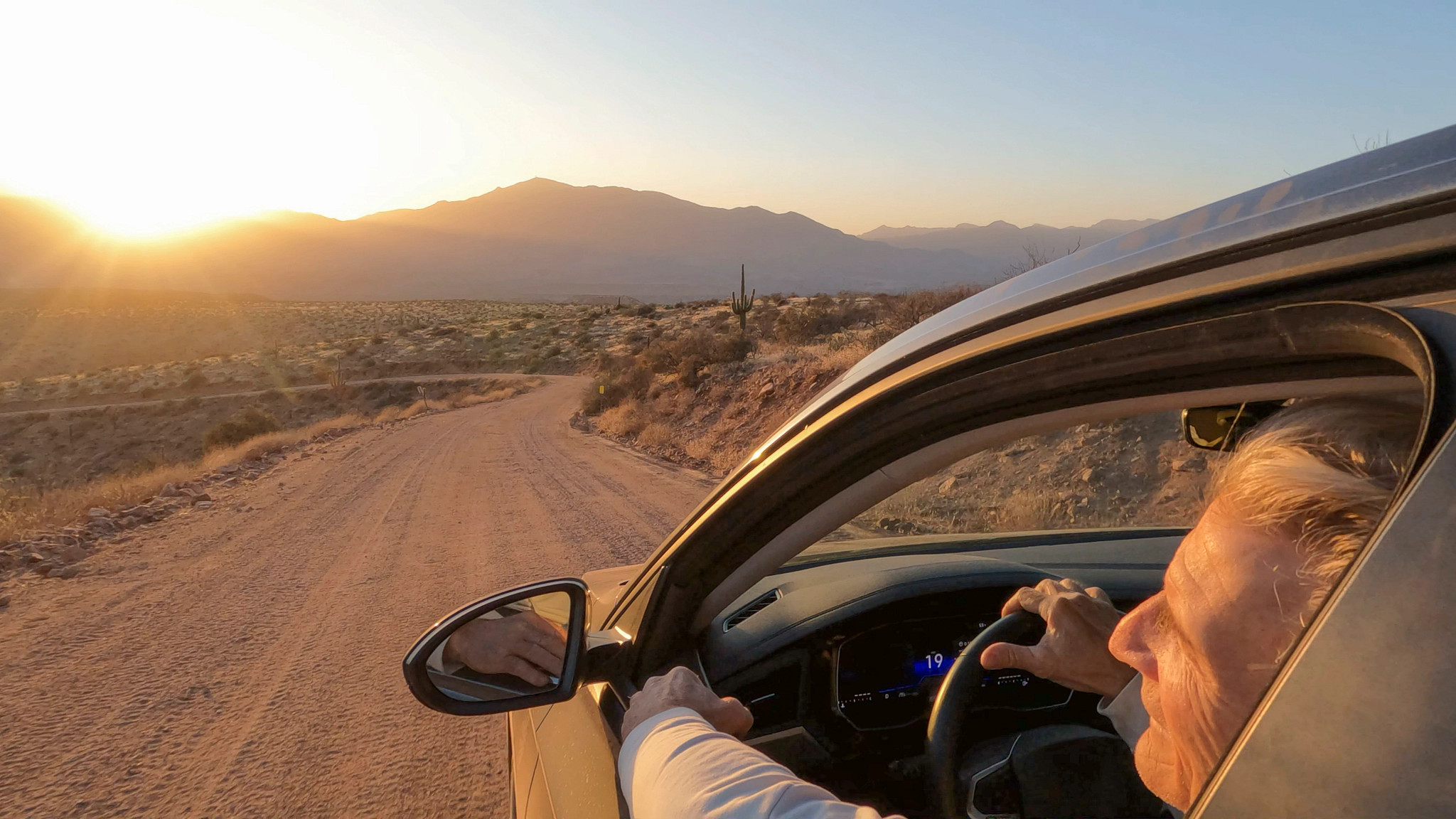 a person looking out the window of a car that's traveling along a dirt road in the desert