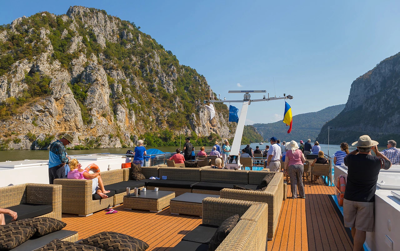 Passengers relaxing on a river cruise ship deck while sailing past dramatic mountain cliffs