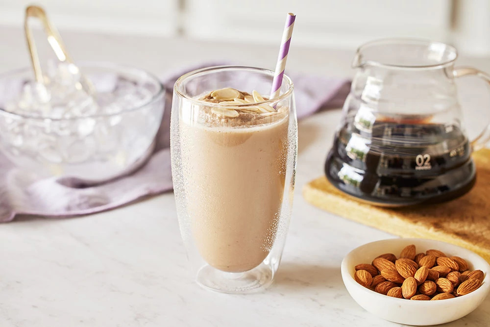Creamy almond smoothie in a double-walled glass with a striped straw beside a bowl of raw almonds