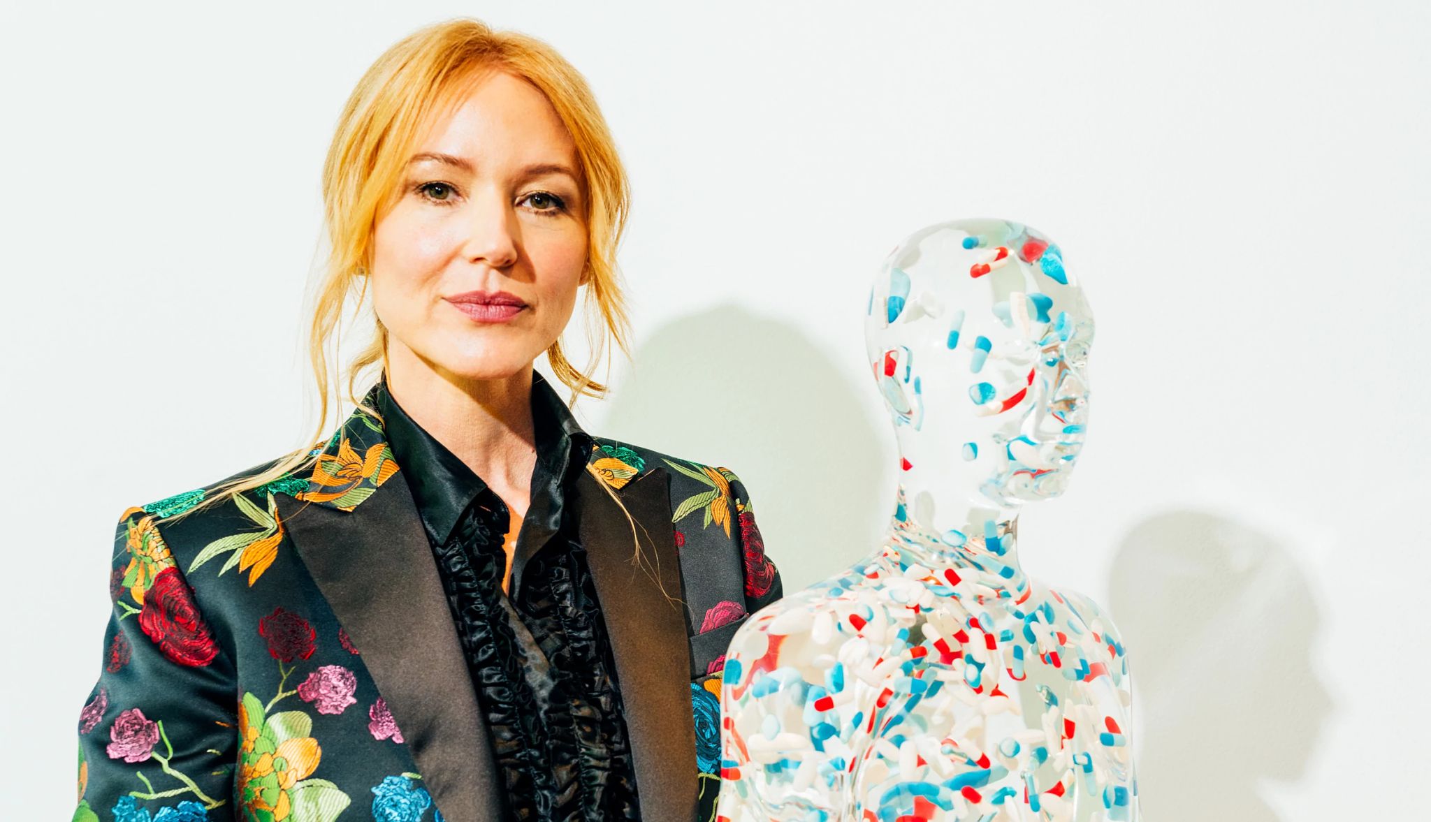 Jewel's sculpture "Chill" is on display Jewel wearing blazer with colorful flowers on it, standing next to sculpture of person with pills all over it