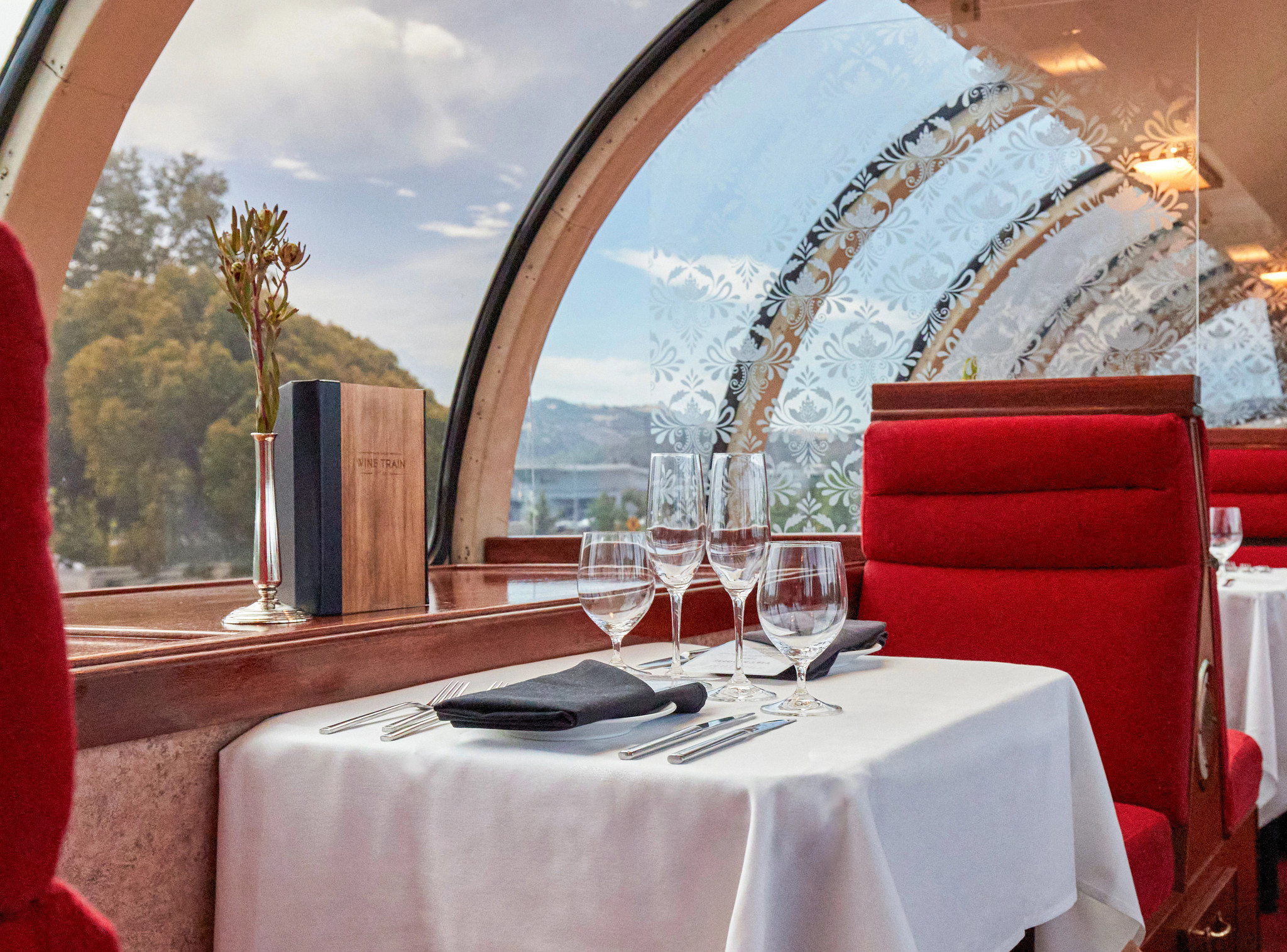 Elegant dining table set inside a scenic wine train with white tablecloth, wine glasses, and red upholstered seating