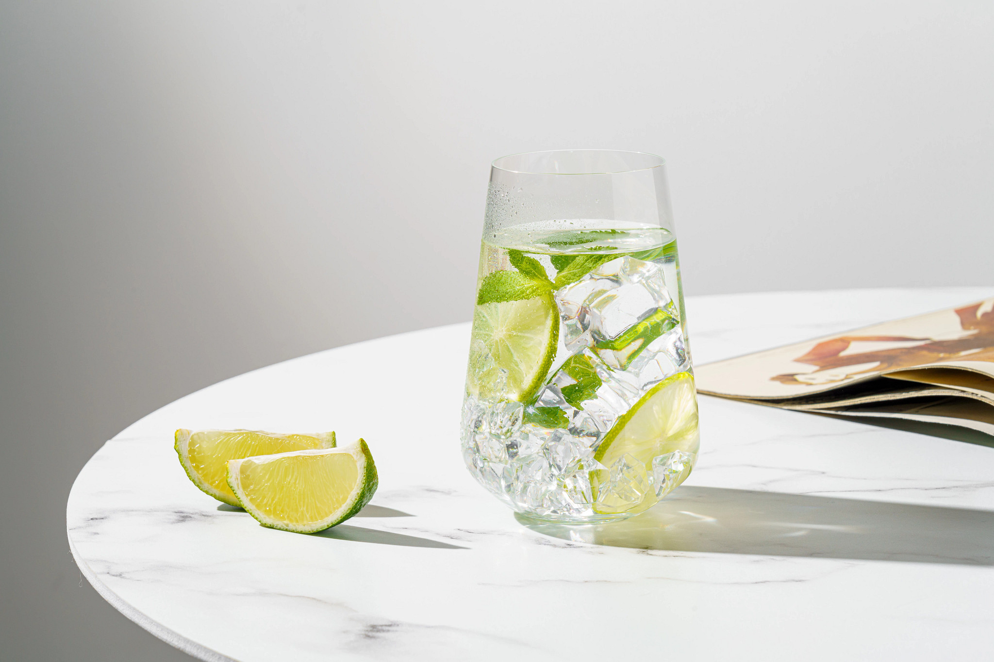 A glass of ice water or sparkling water garnished with fresh lime slices and mint leaves, sitting on a white marble table next to extra lime wedges.