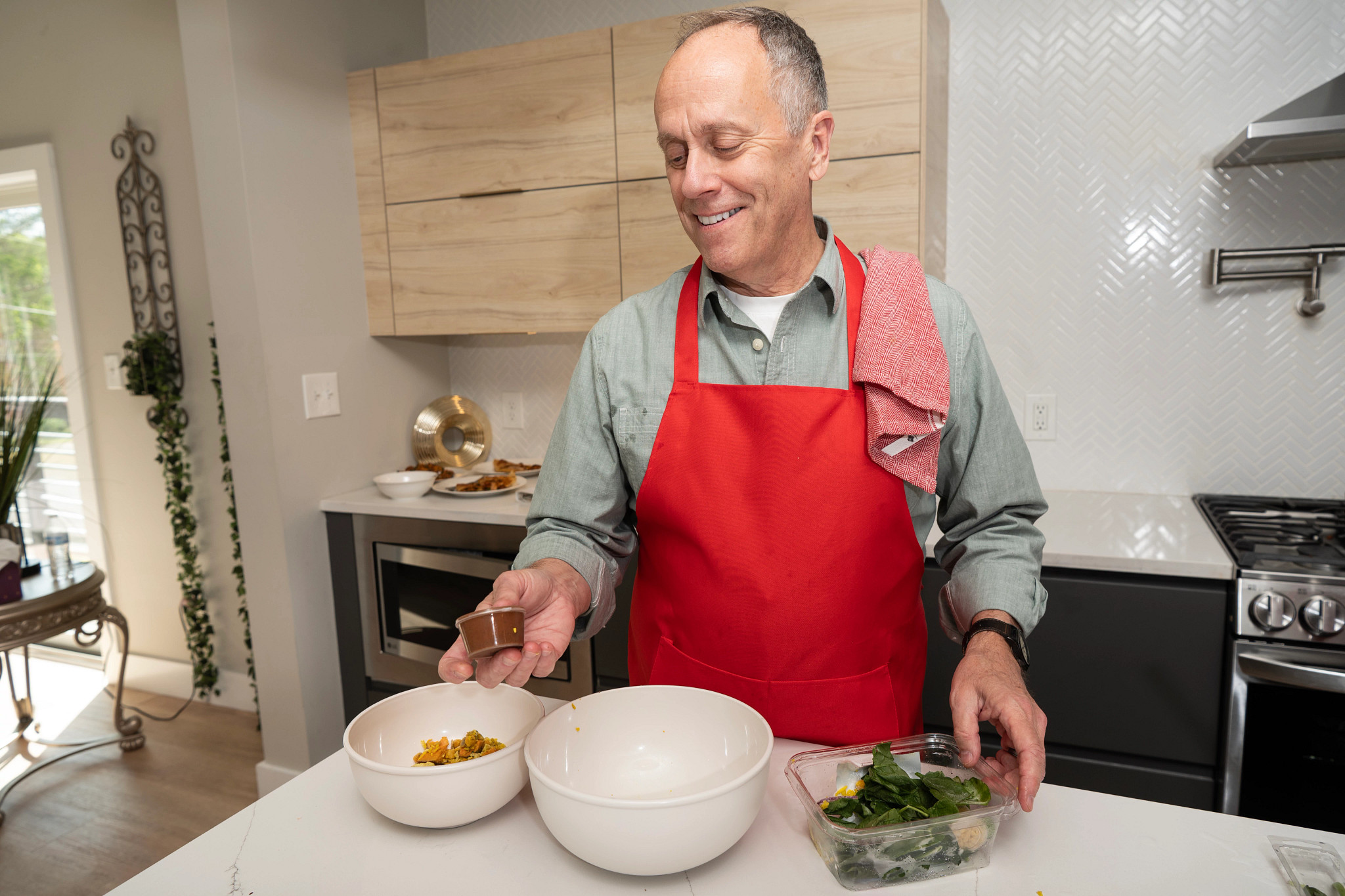 A man in a red apron in a kitchen holding a dressing package with a salad and two food bowls in front of him