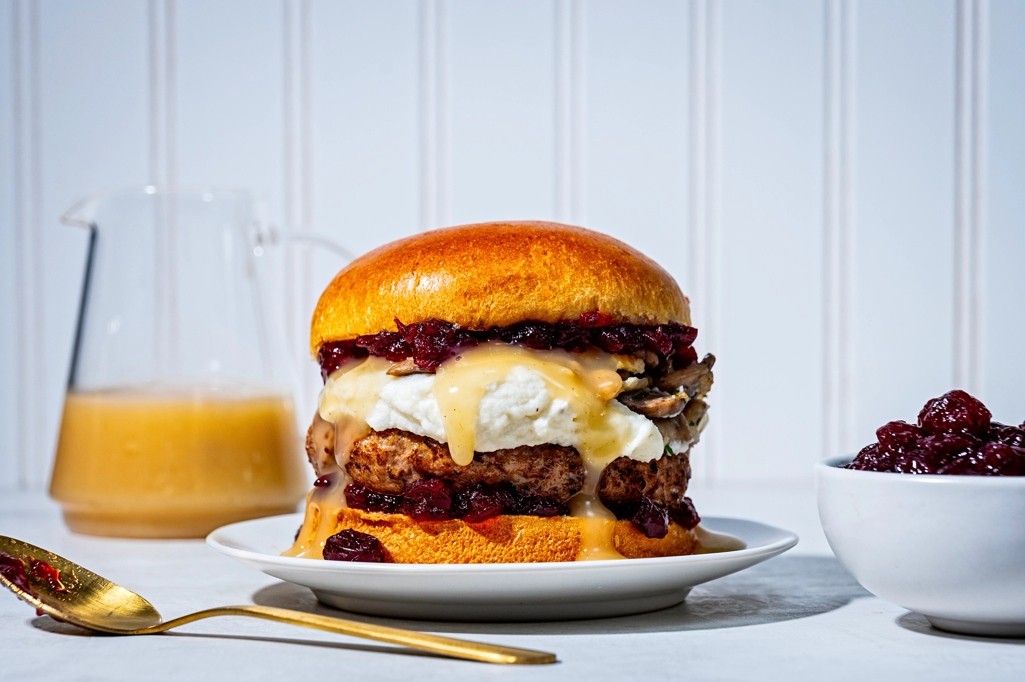 a photo shows a thanksgiving leftover turkey burger is shown on a small white plate. The burger is oozing with mashed potatoes, cranberry sauce and turkey gravy