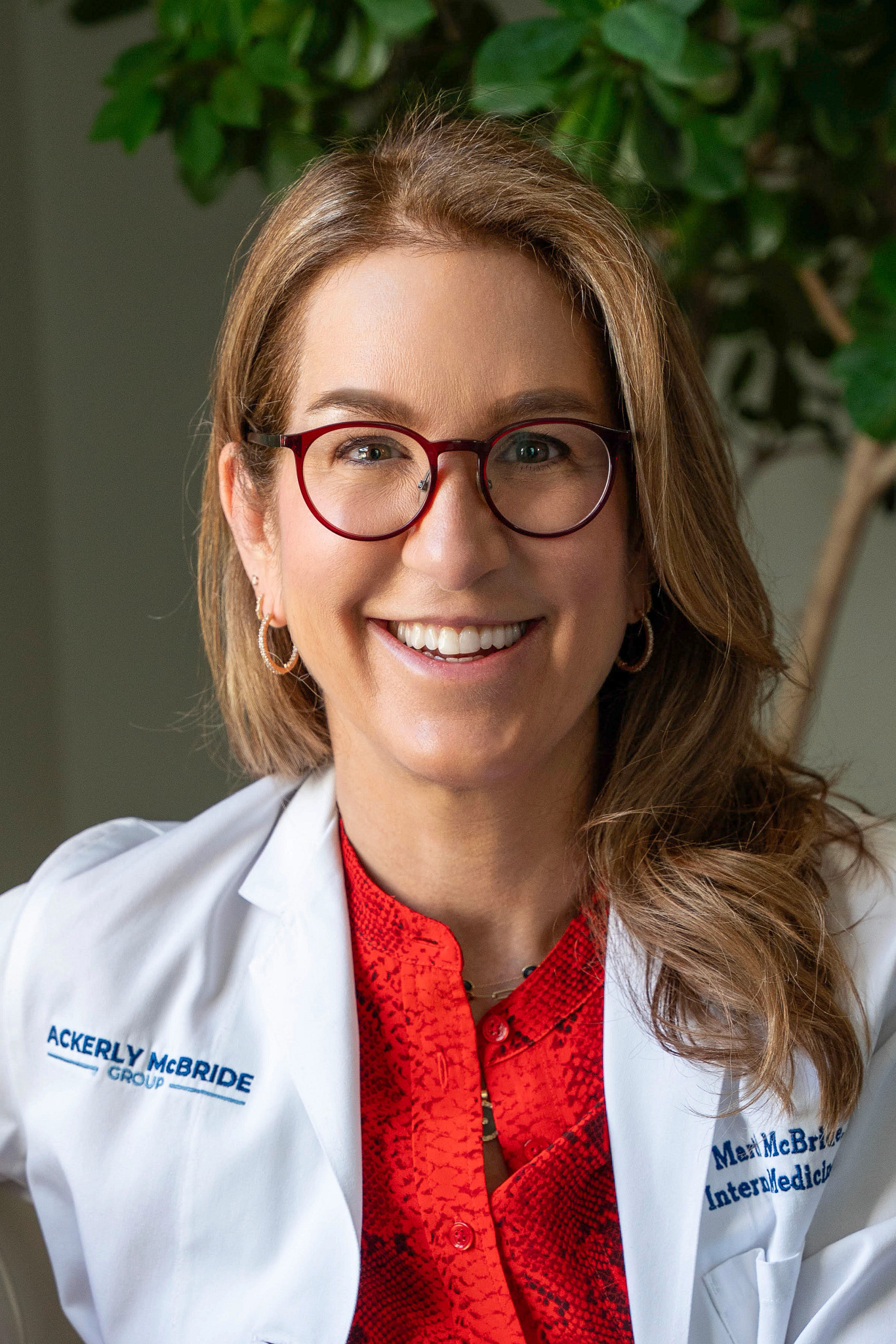 Headshot of Dr. Lucy McBride wearing glasses, a red shirt, and a white lab coat while smiling.