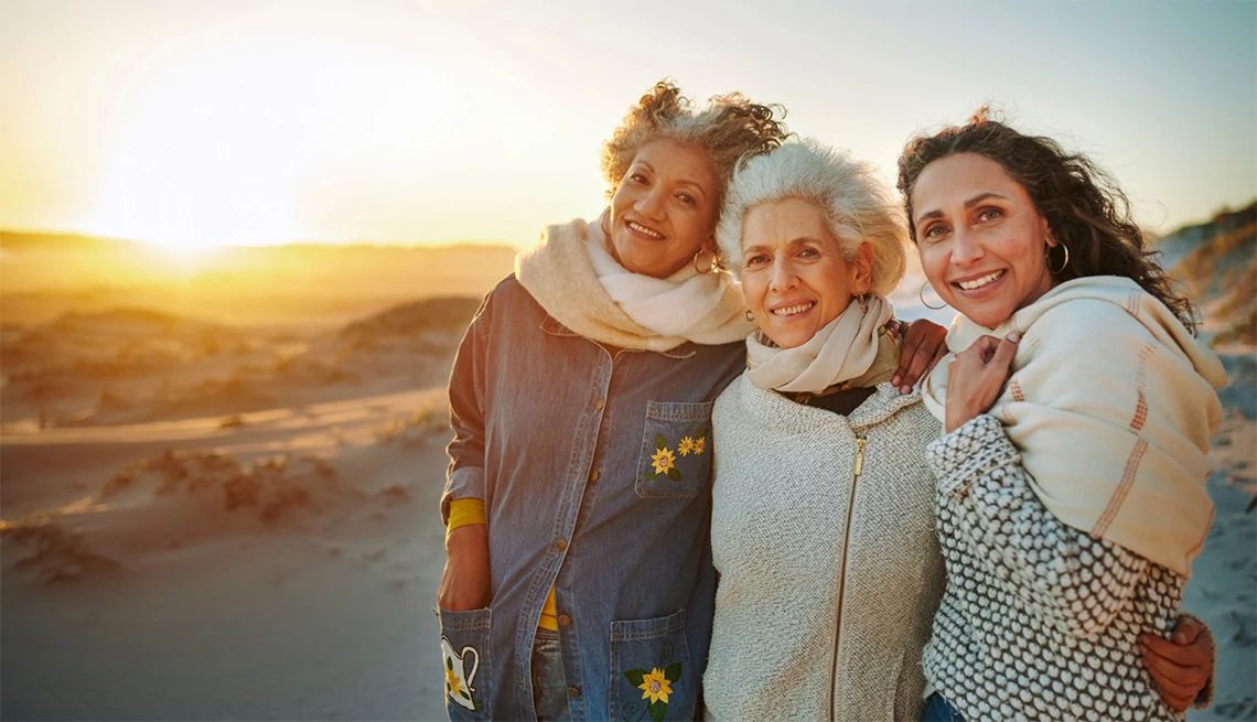 AARP Navigator Maximizing Retirement Income Three women standing on a beach at sunrise wearing coats as their hiar blows in the wind