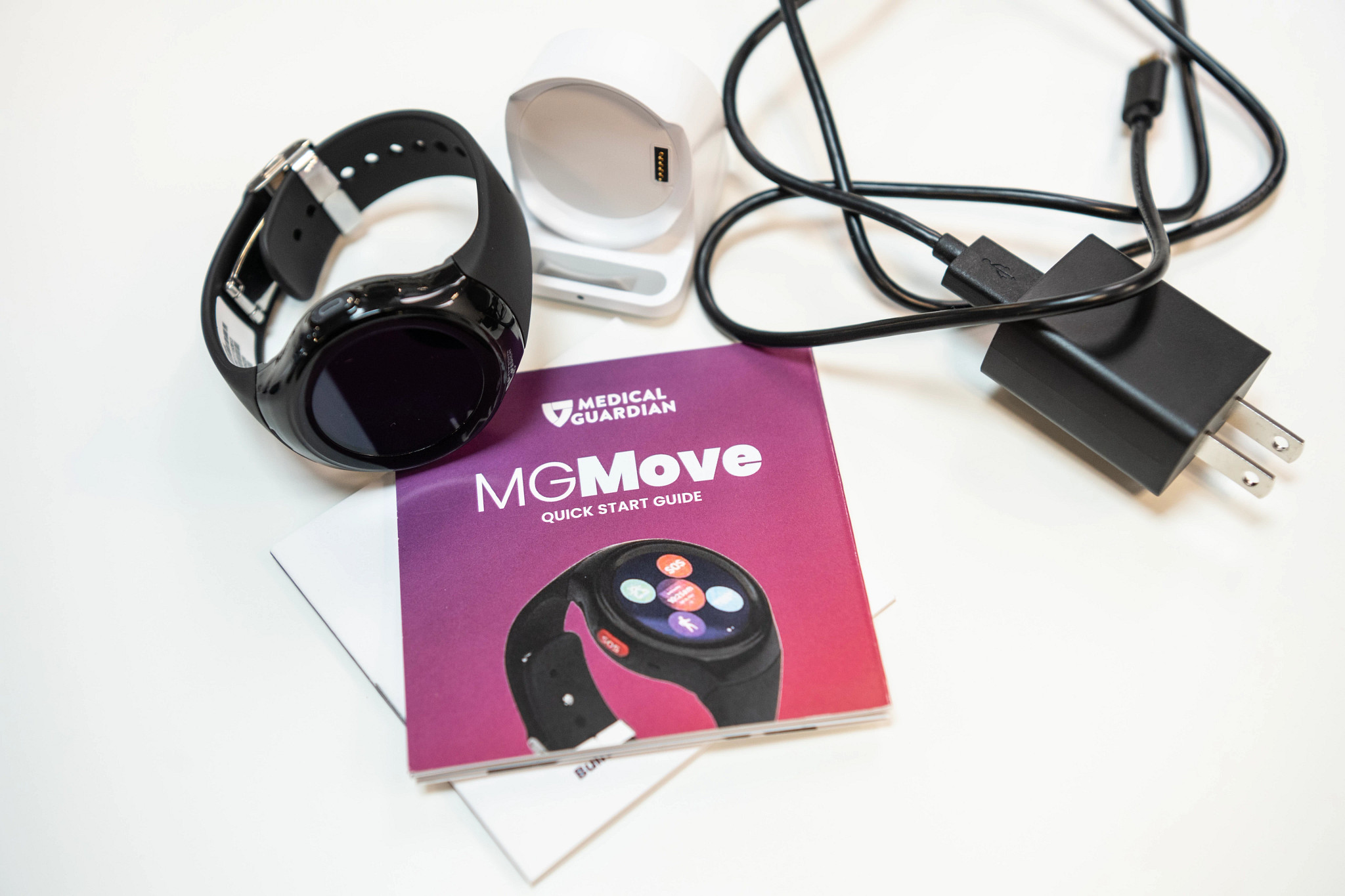The MGMove smartwatch medical alert system next to its charging port, charging cable, and user manual on a white table