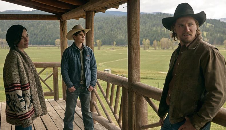 Scene from Yellowstone A man, a woman and a teen boy standing on a front porch with mountains in the background