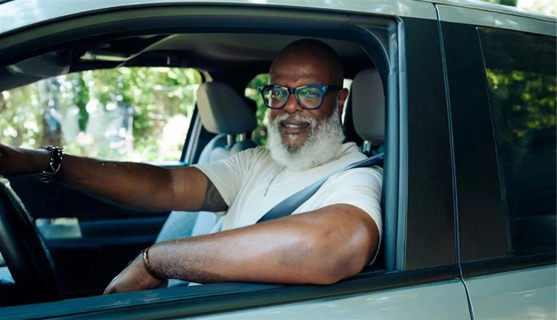 Mature man with a grey beard and eye glass leaning his arm on the window of his car while sitting in the driver's seat