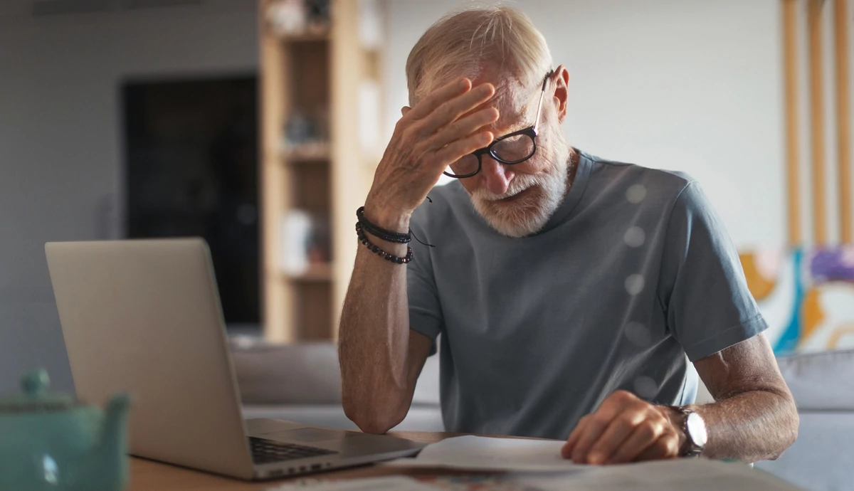 An older man looking at medical bills with worry while sitting at a table with his laptop