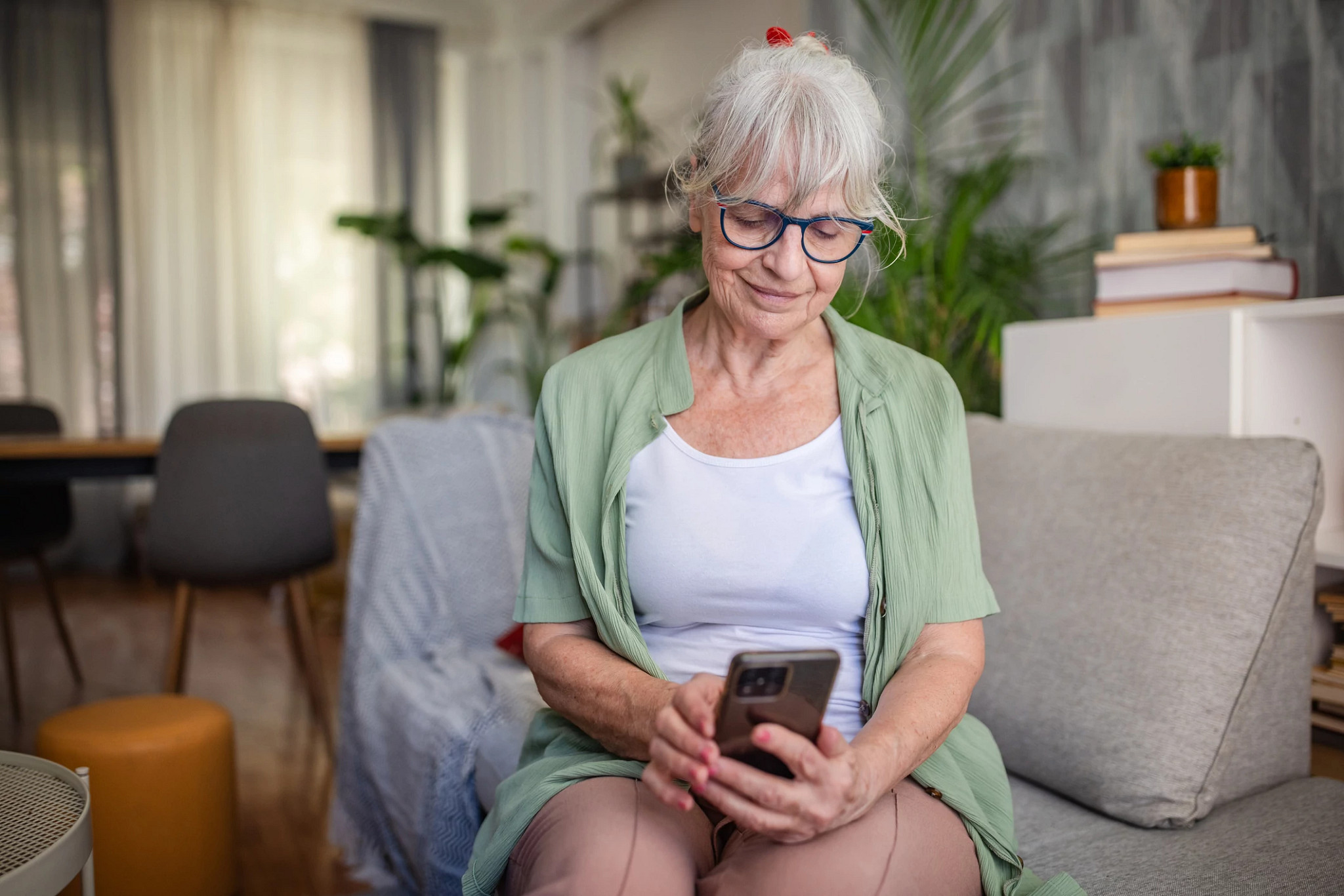 An elderly woman in a green cardigan and white top smiles while engaging with her smartphone, seated comfortably on a couch amidst homey furnishings and plants around.