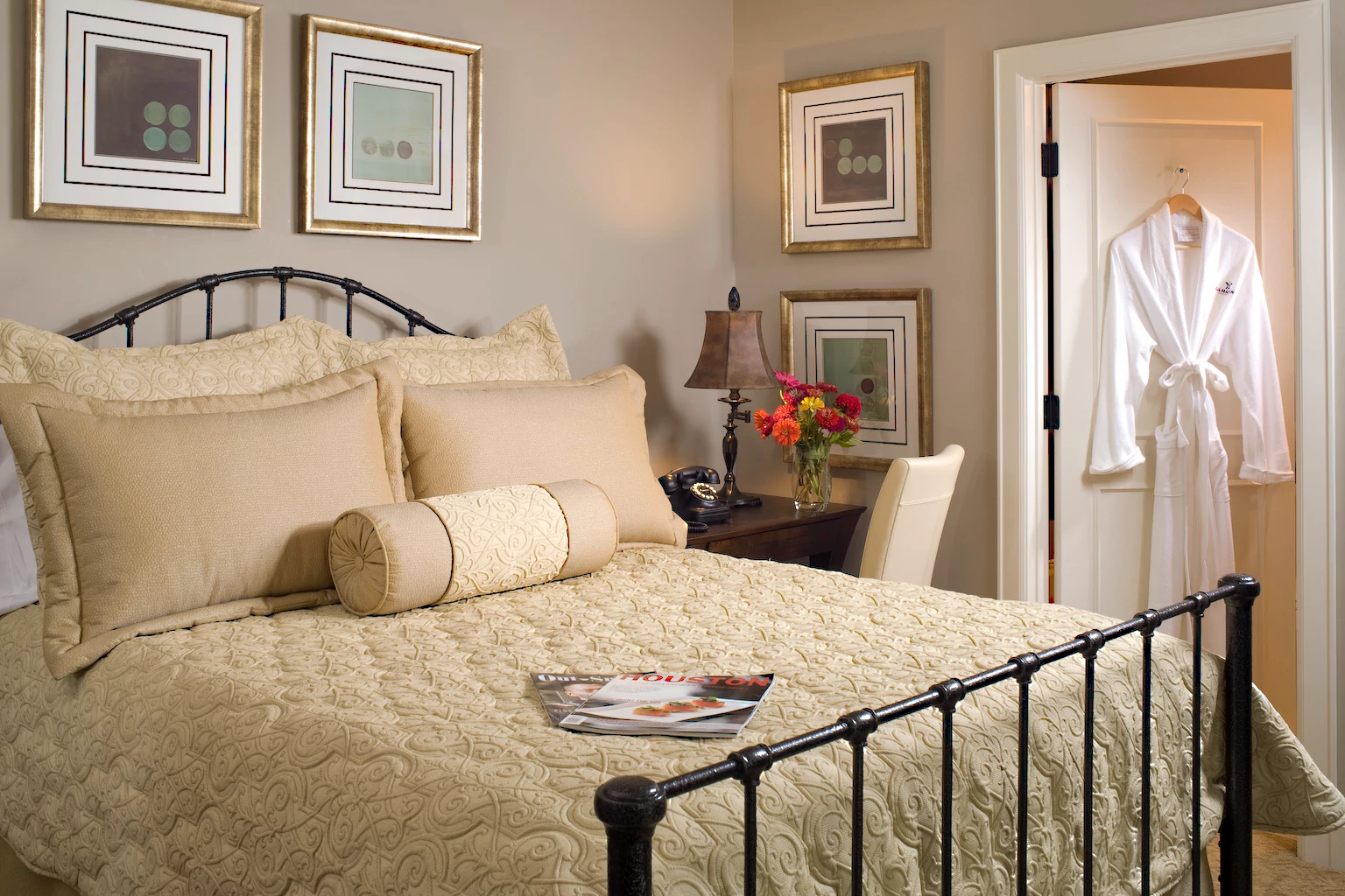 a hotel room with cream bedding, art on the walls and a white robe hanging on a door