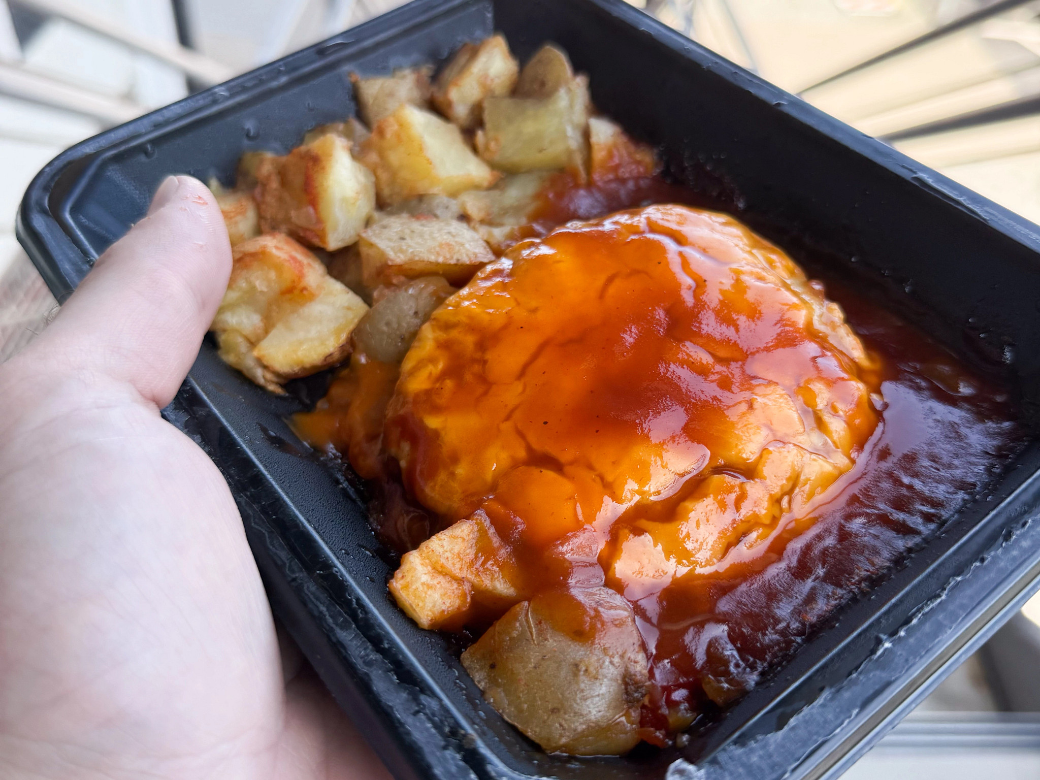 A person’s hand holding the edge of a black container filled with diced potatoes and sauce over cheese