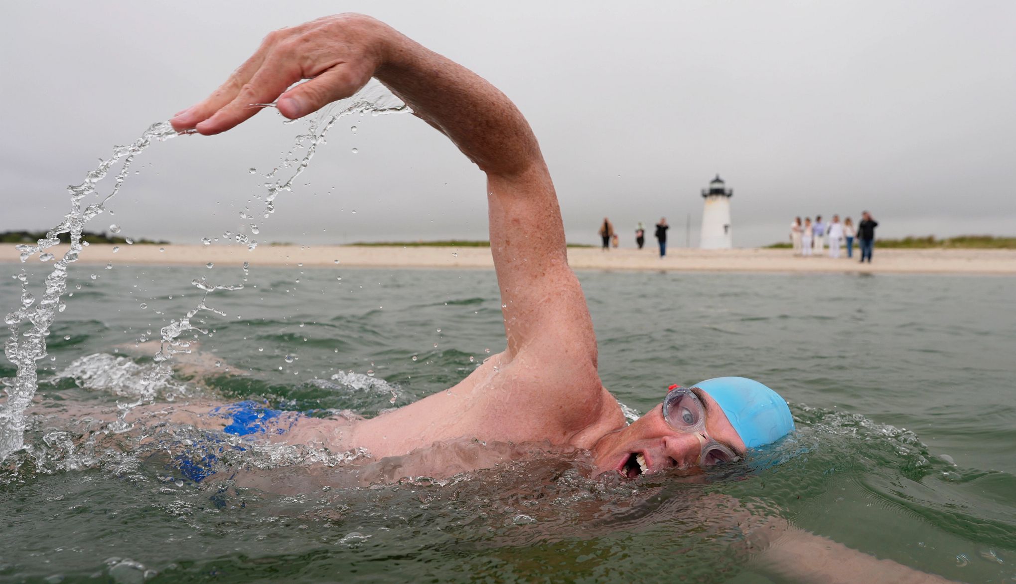 Martha’s Vineyard Swimmer Endurance swimmer Lewis Pugh is shown in a photo swimming near the Edgartown Harbor Light in Edgartown, Mass. There are people on the beach and a lighthouse in the distance.