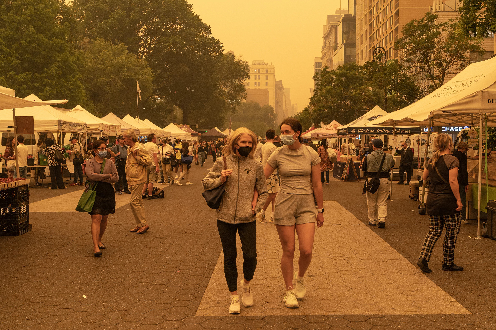 An older woman and a young woman wearing face masks walk through an outdoor market under a thick, orange haze caused by wildfire smoke.