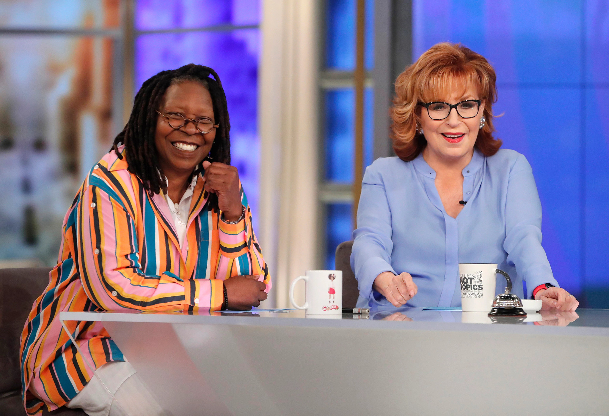 Whoopi Goldberg sitting next to Joy Behar at a desk with a mug in front of each of them