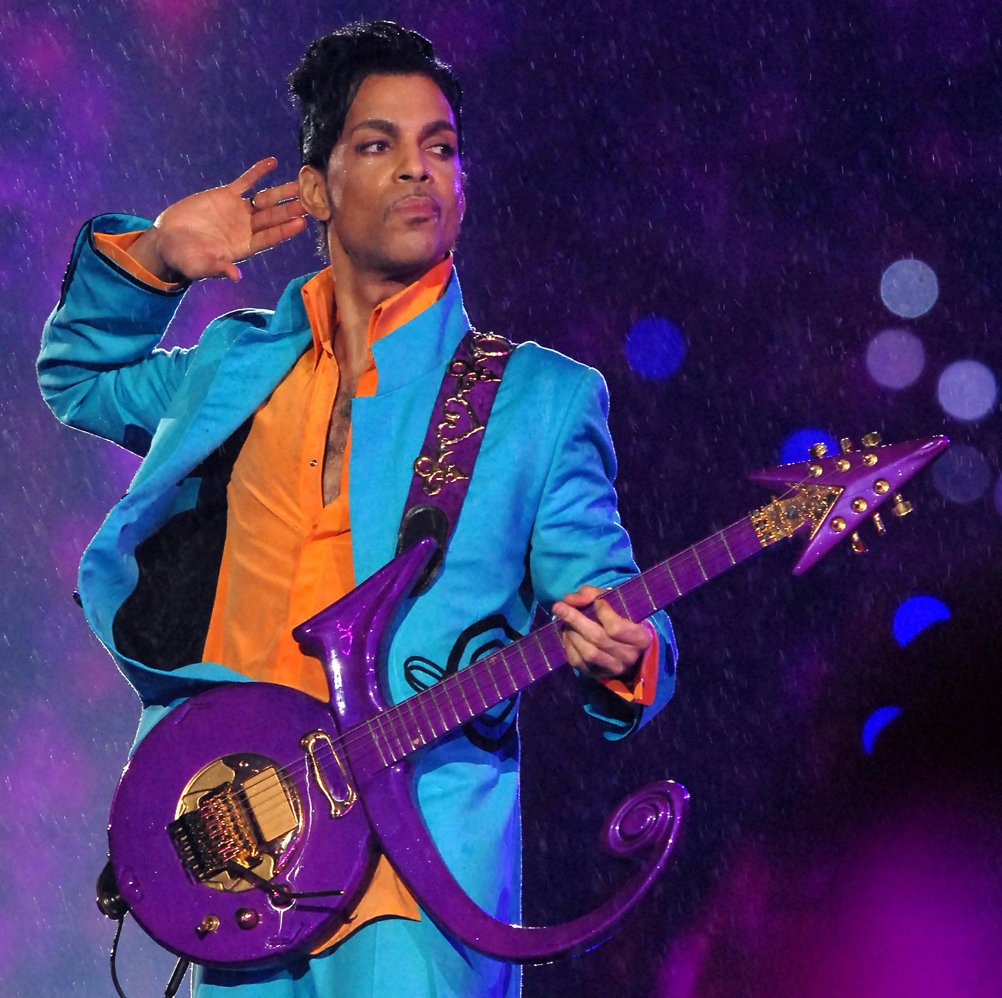 Prince posing with his right hand to his ear and a serious expression while performing with a guitar onstage