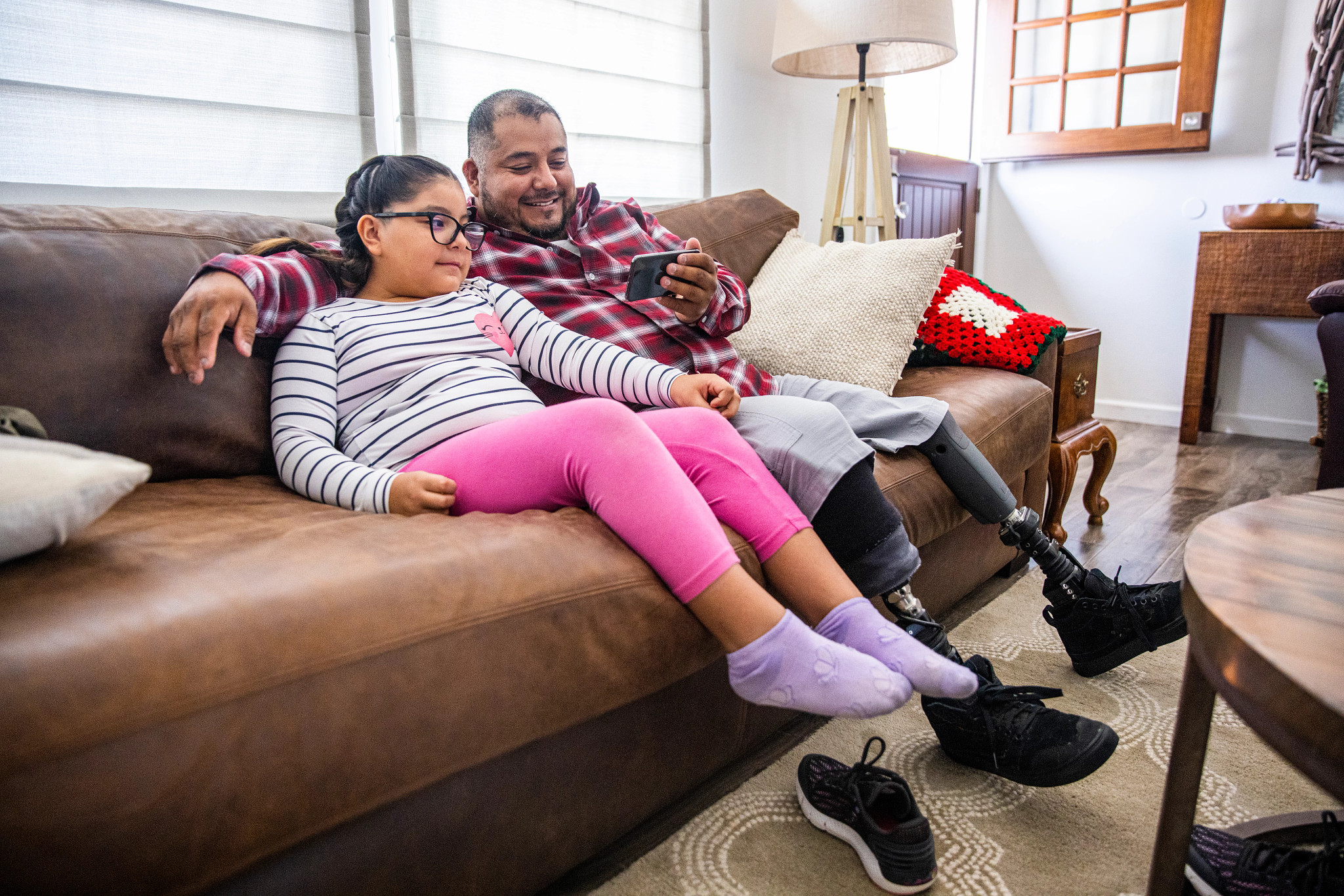 a man with prosthetic legs sits on a couch with a child, watching something on his smartphone