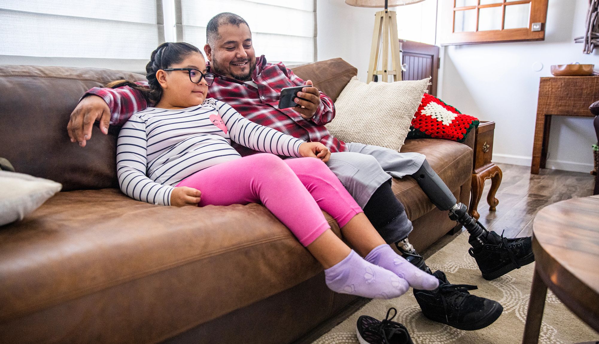 a man with prosthetic legs sits on a couch with a child, watching something on his smartphone