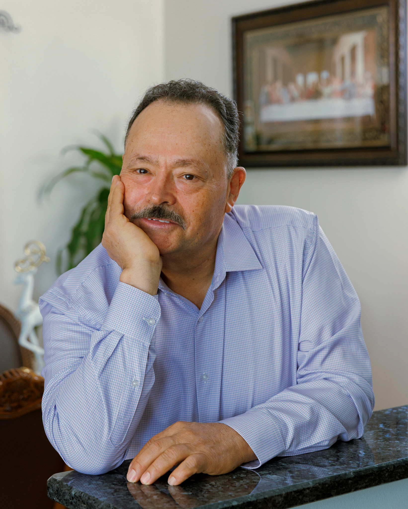 portrait of Elpidio Gutierrez, a man with a mustache wearing a light blue checked shirt, leaning forward thoughtfully with his hand on his chin