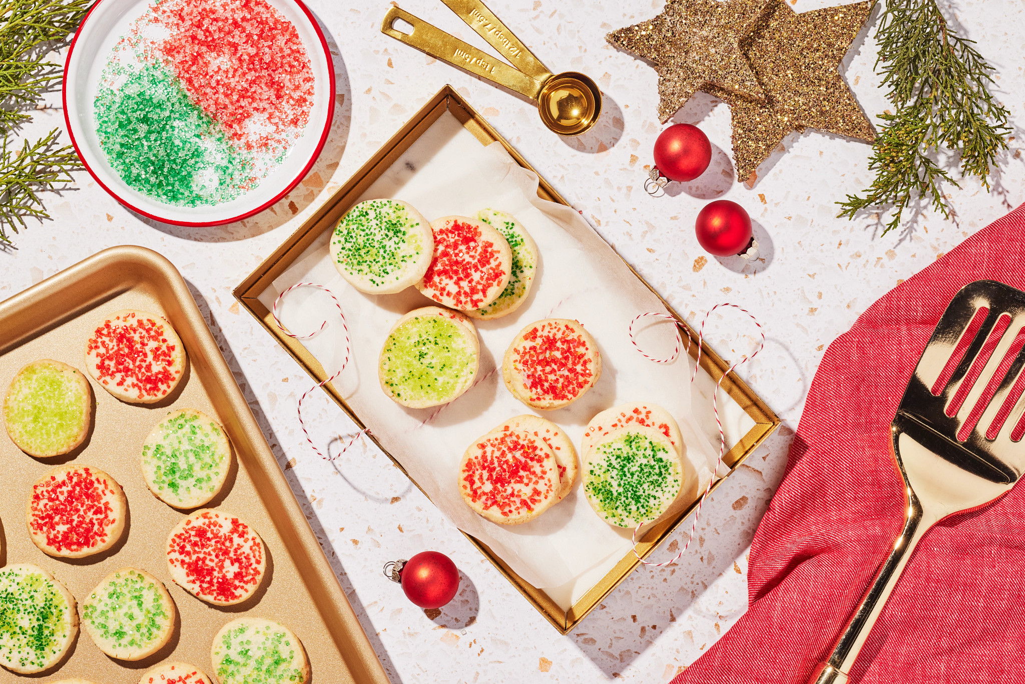 Frosted Christmas cookies with red and green sprinkles arranged on a baking sheet beside a holiday gift box