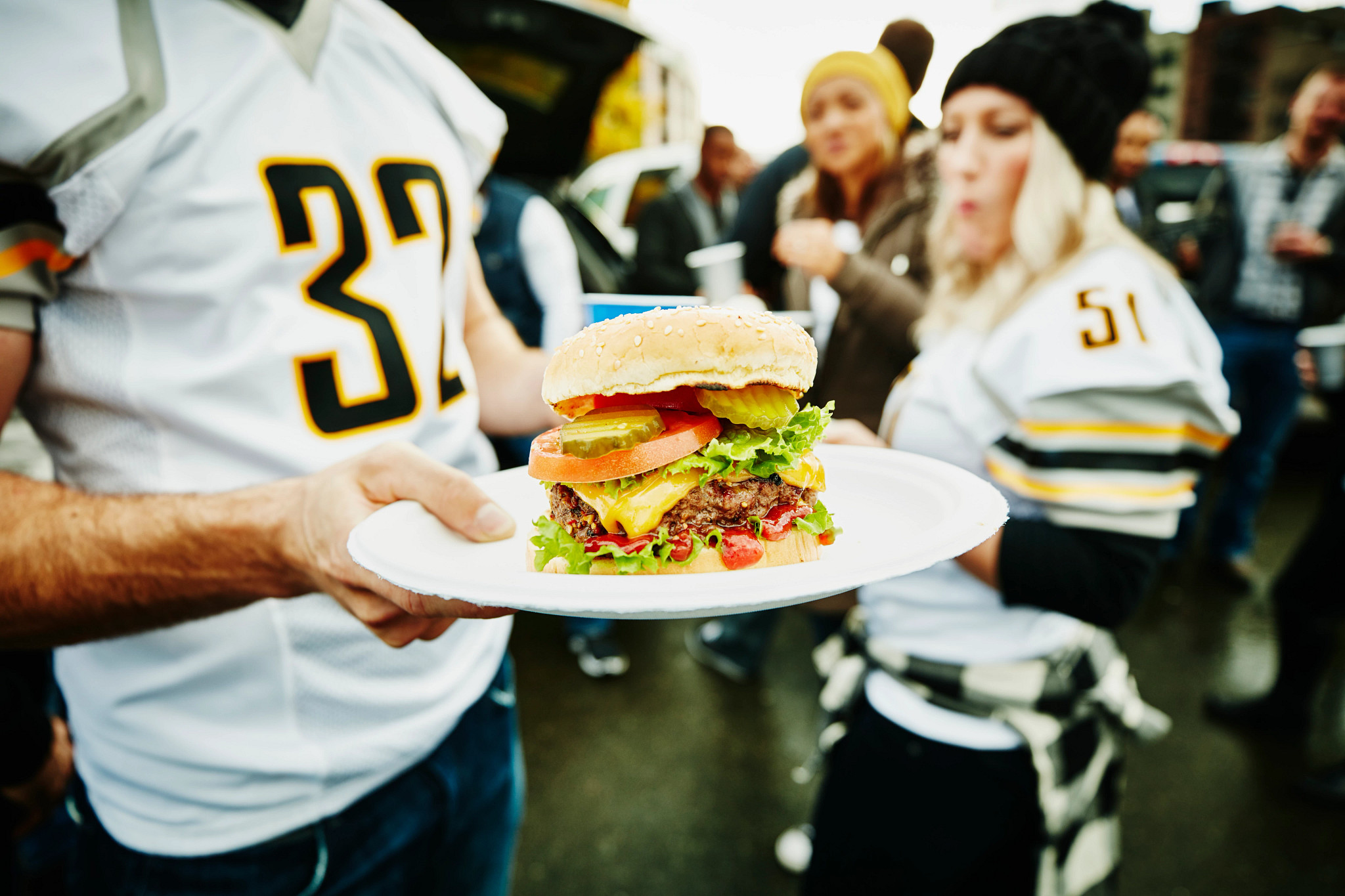 A photo shows a tailgater brandishing a tasty burger while other revelers look on in admiration