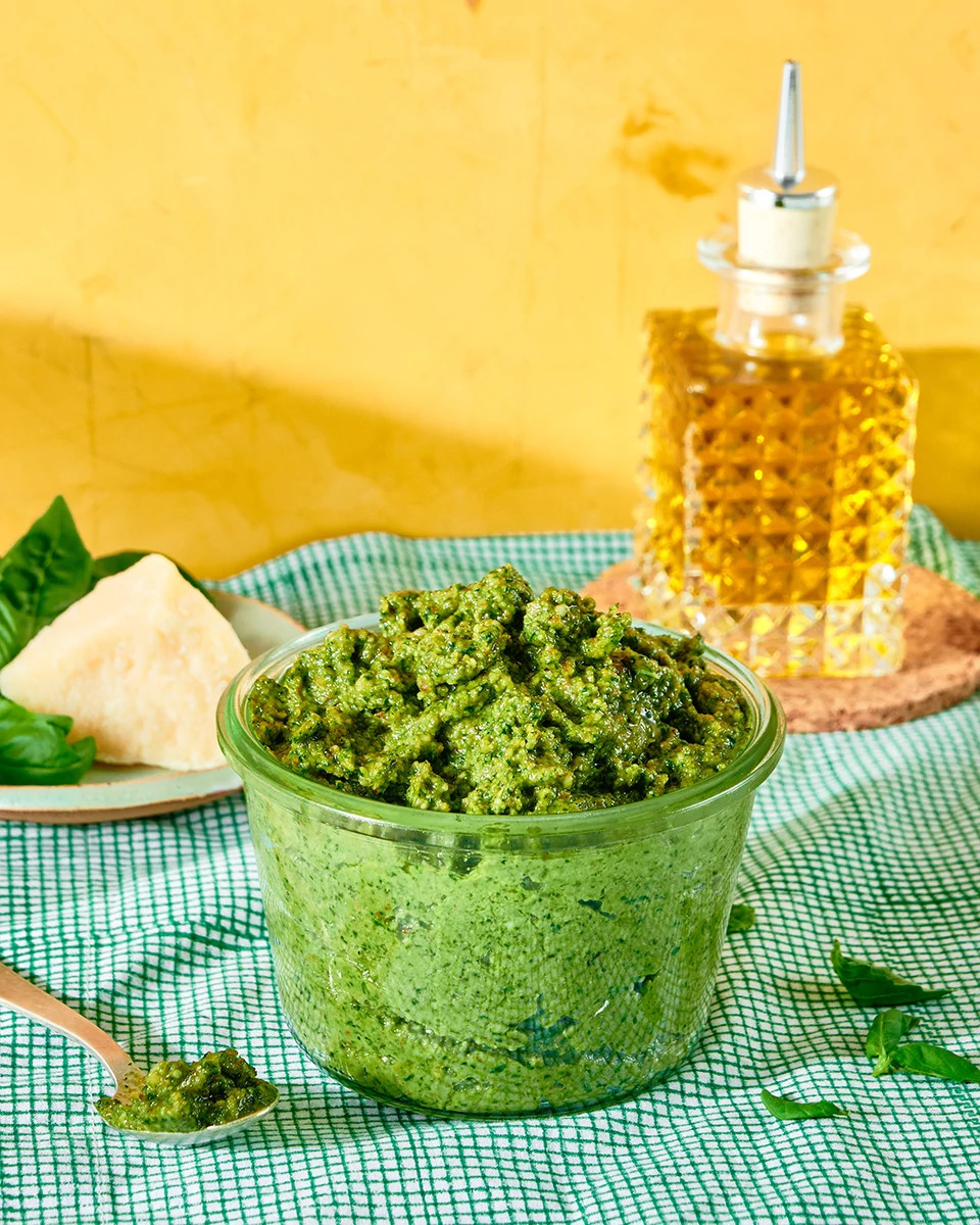 Fresh basil pesto in a glass bowl with parmesan cheese and olive oil on a checkered kitchen towel